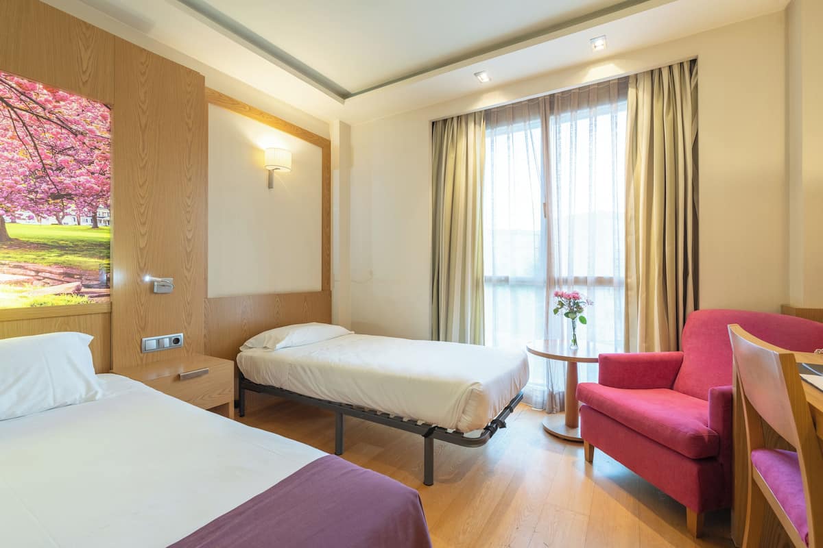 Quarto de hotel com duas camas, uma poltrona vermelha, uma mesinha e um quadro de uma cerejeira.