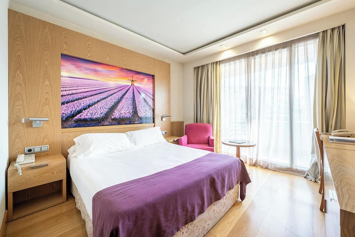 Quarto de hotel com uma cama grande, uma mesa de cabeceira, uma escrivaninha, uma cadeira e um quadro de um campo com uma cruz.