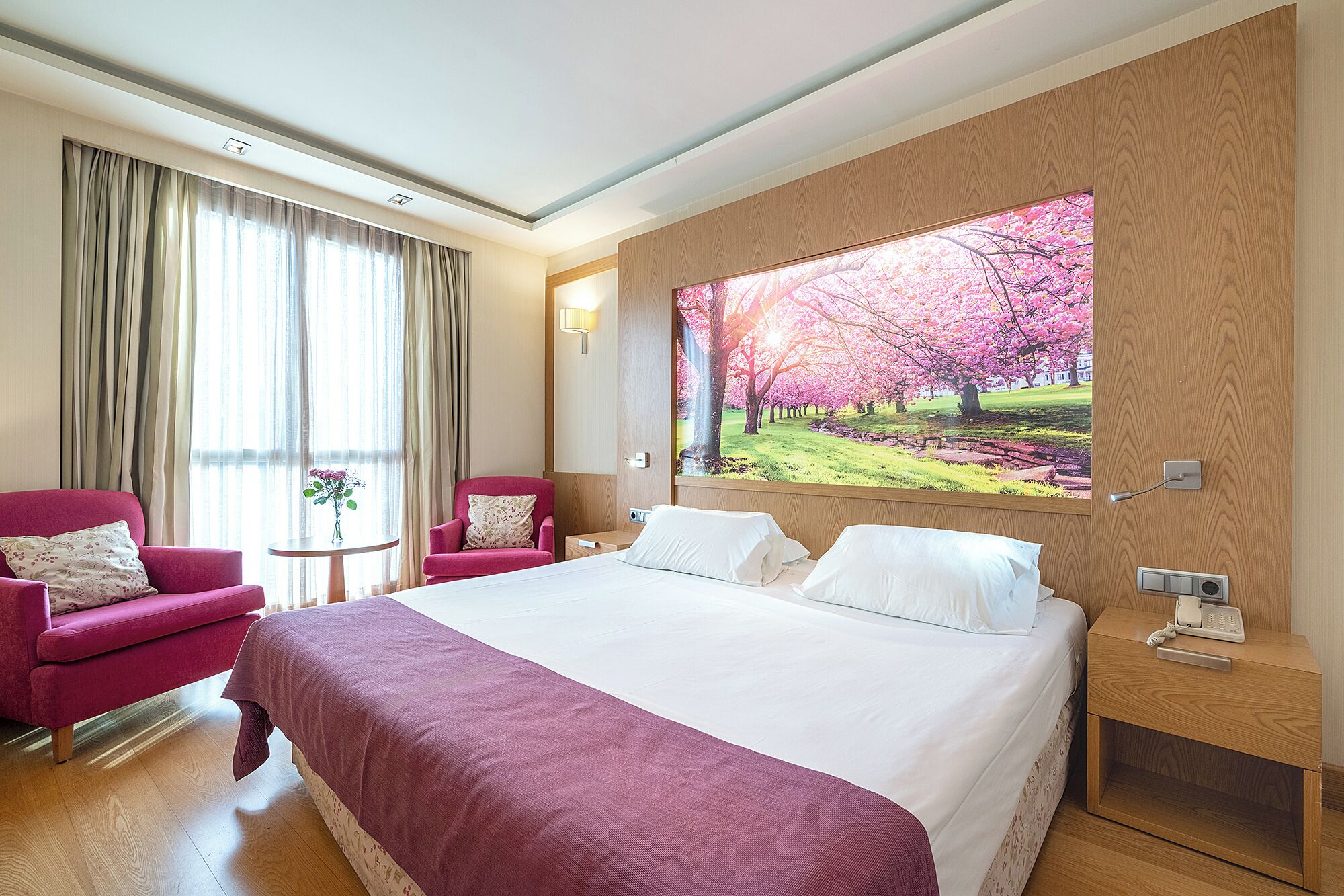 A hotel room with a large bed, two armchairs, a small table with a vase, and a wall-mounted TV displaying a nature scene.