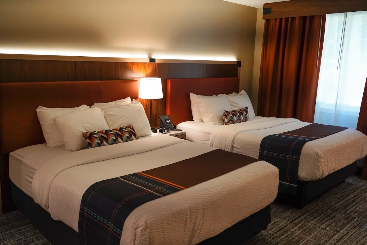 Une chambre d’hôtel avec deux lits, chacun agrémenté de draps blancs et d’un plaid marron. On y trouve une table de chevet avec une lampe et un téléphone. La chambre est dotée d’une tête de lit en bois et d’une fenêtre avec des rideaux.