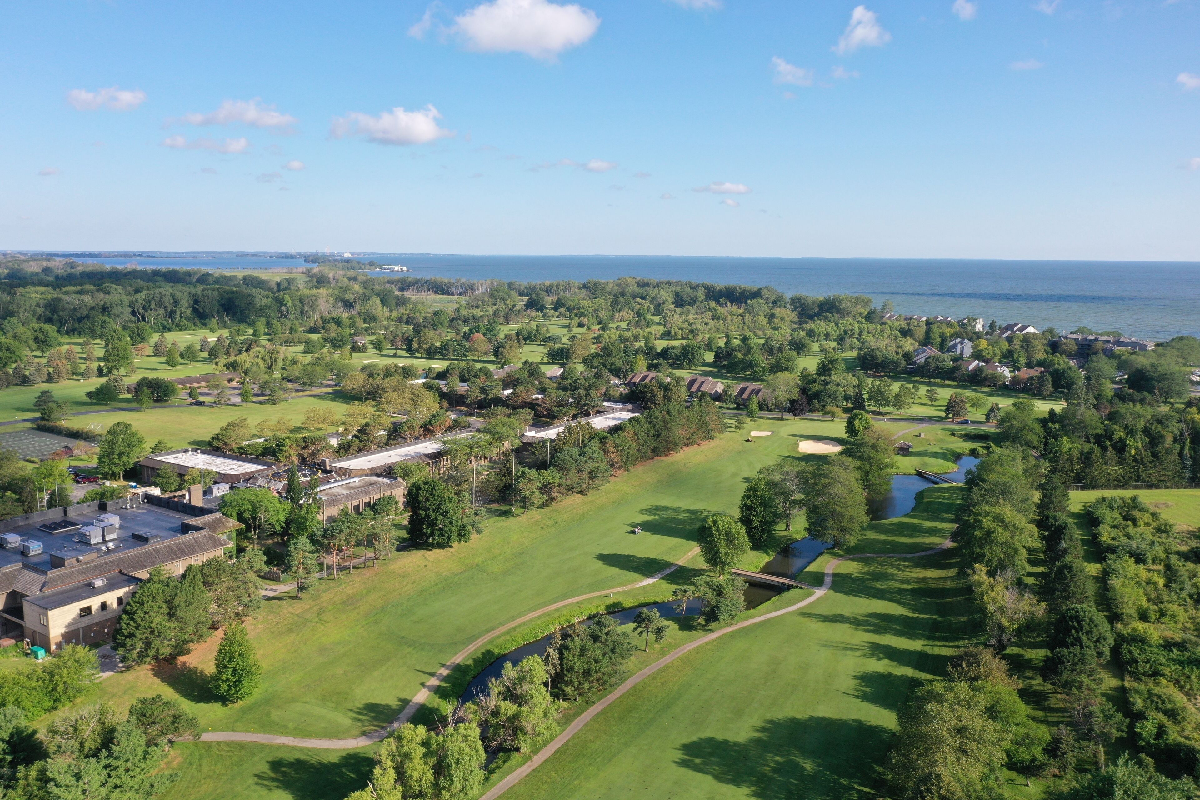 An aerial view of a golf course with a clubhouse, surrounded by trees and a body of water.