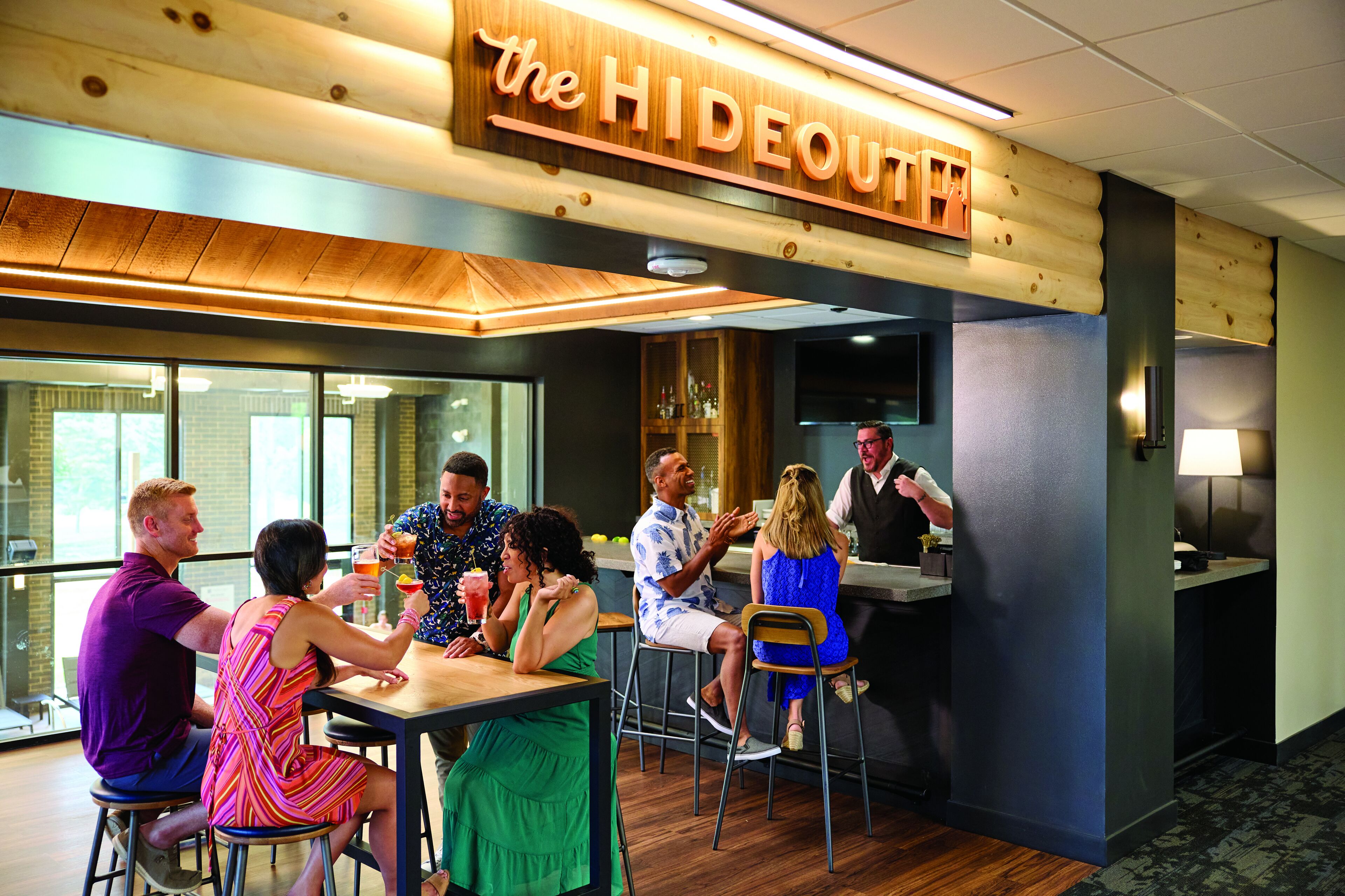A group of people seated at a bar area with a wooden ceiling and a sign that reads "the HIDEOUT".