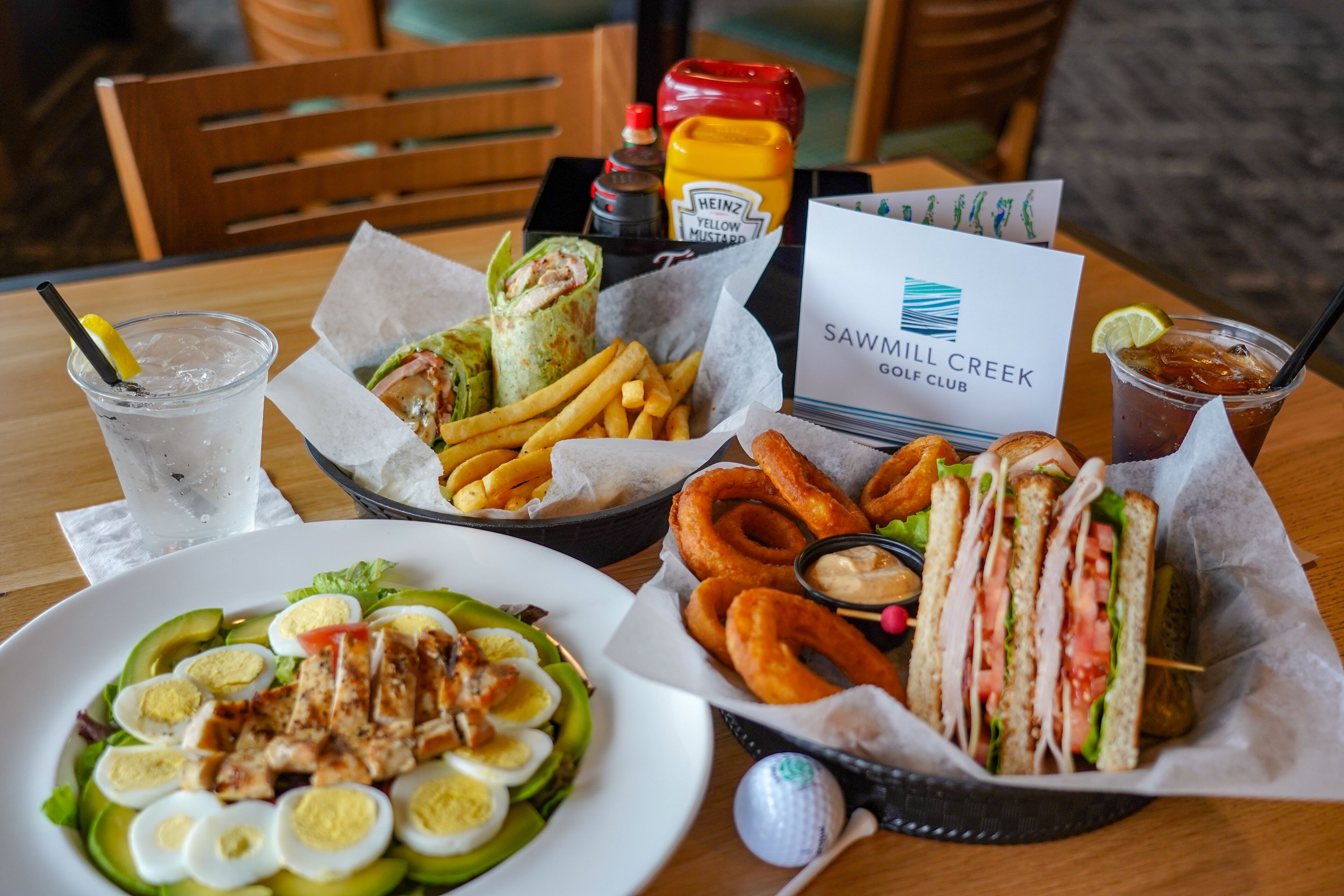 A table with a variety of food items including a salad, sandwiches, fries, and a drink. There is a sign for Sawmill Creek Golf Club.