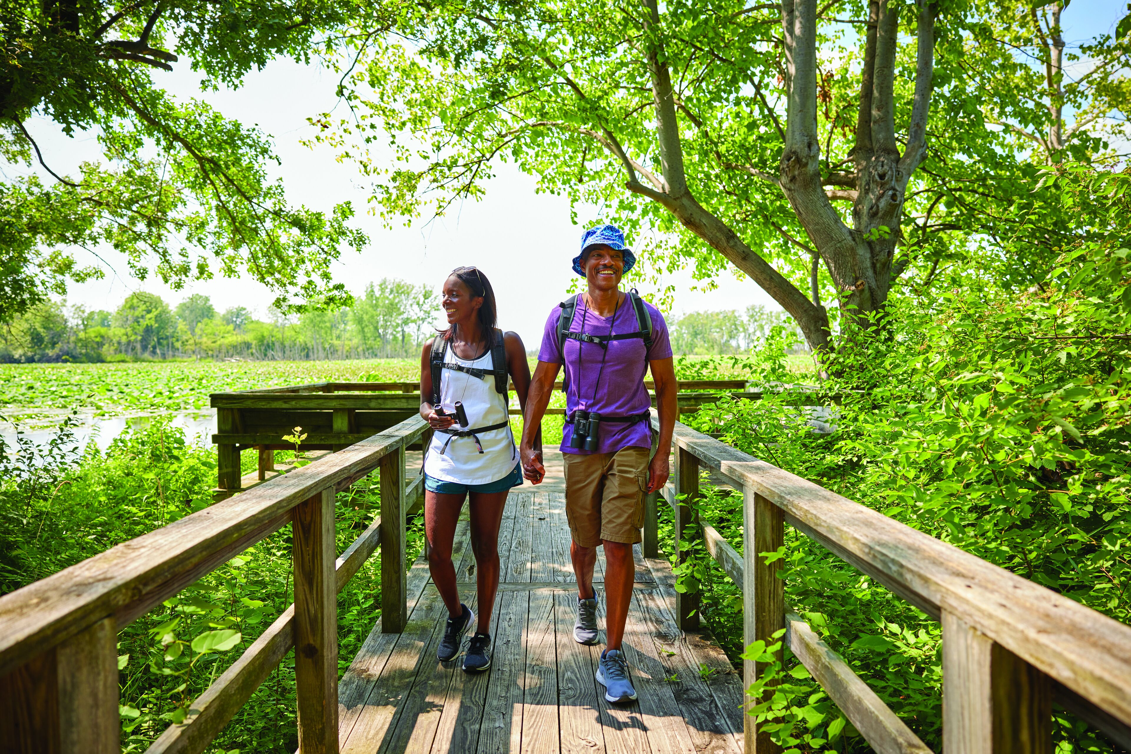 Two people walking on a wooden bridge surrounded by greenery.
