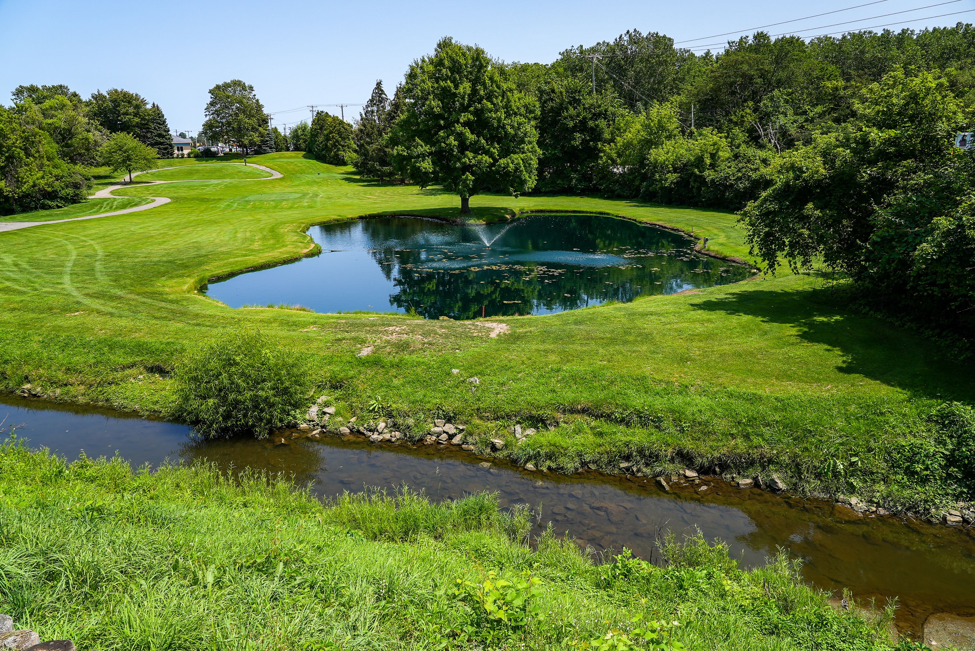 Ein Golfplatz mit Teich, einem kleinen Bach und einem Haus im Hintergrund.