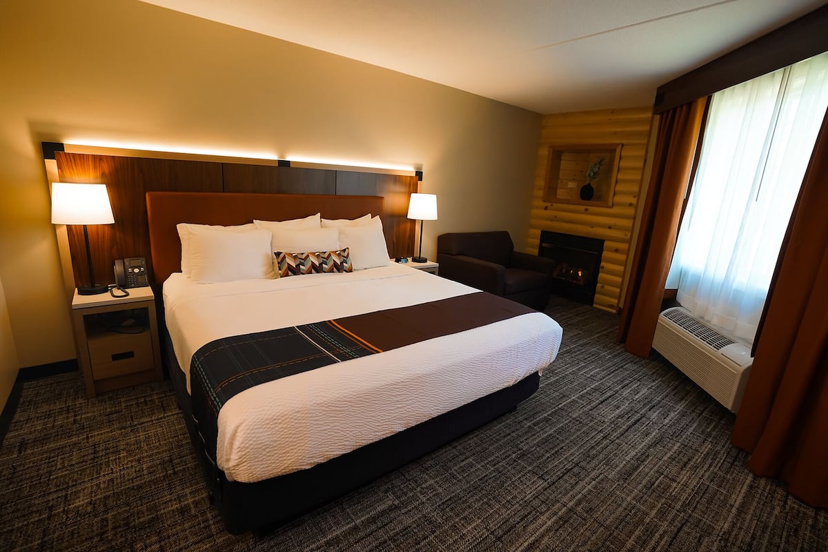 Une chambre d’hôtel avec un grand lit, deux lampes de chevet, un fauteuil, une cheminée et une fenêtre avec des rideaux.