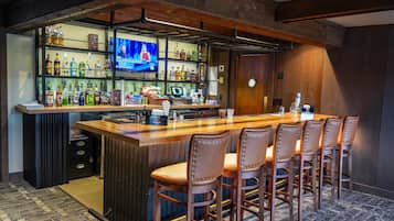 A bar area with a long counter, multiple shelves of bottles, a television screen, and bar stools.