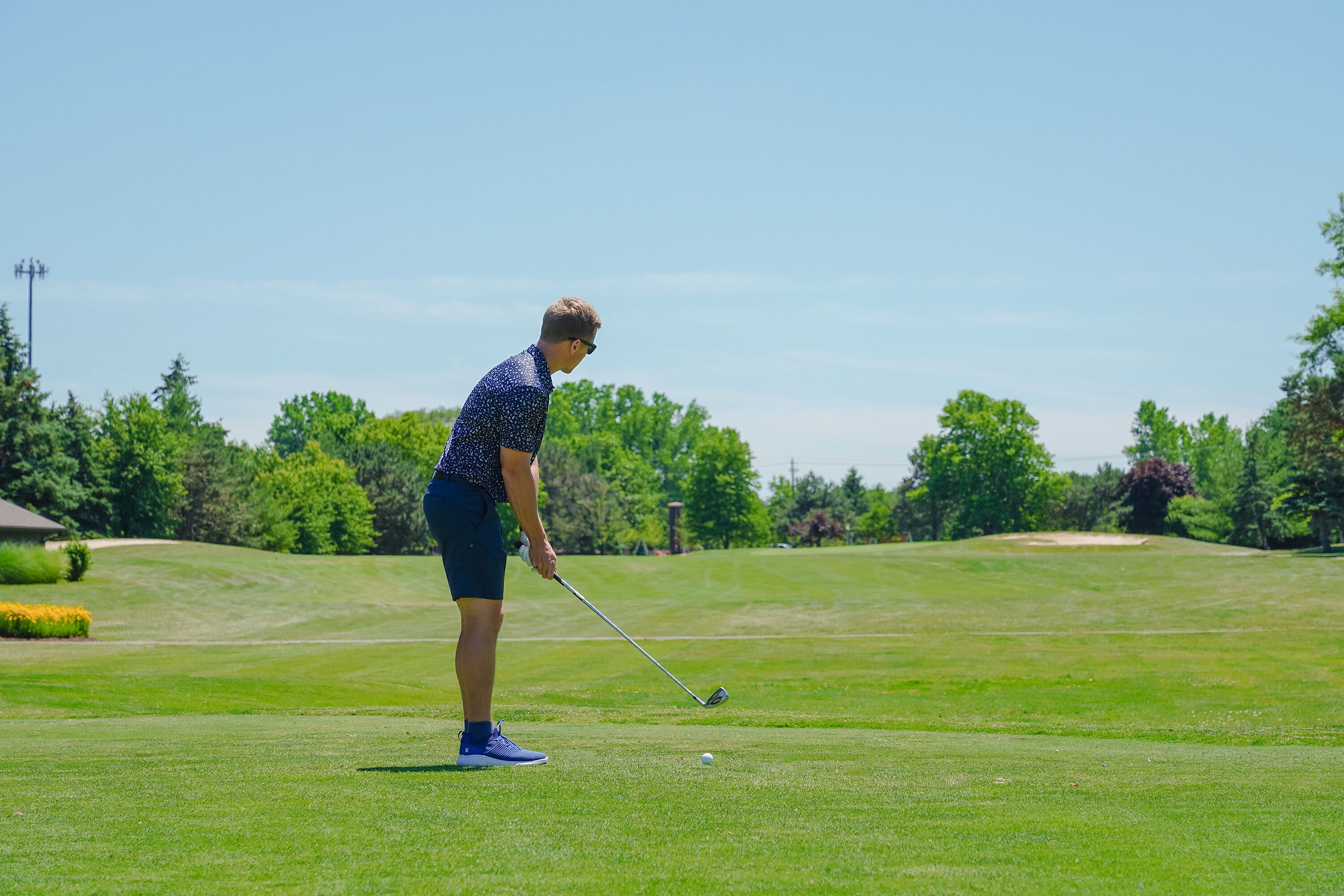 A person playing golf on a well-maintained course with trees and a clear sky in the background.