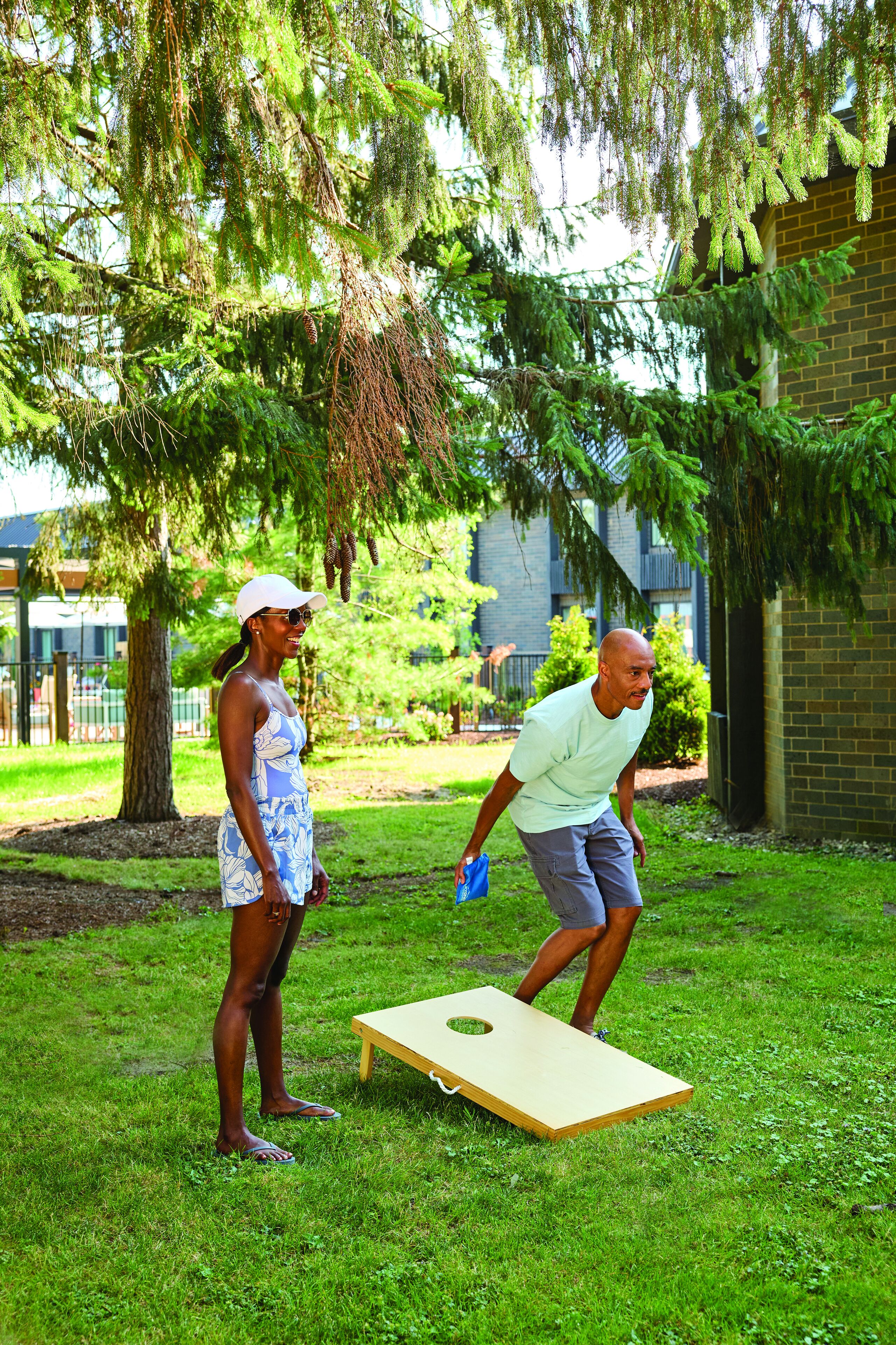 Two people playing a game on a grassy area with a wooden board, surrounded by trees and a brick building.