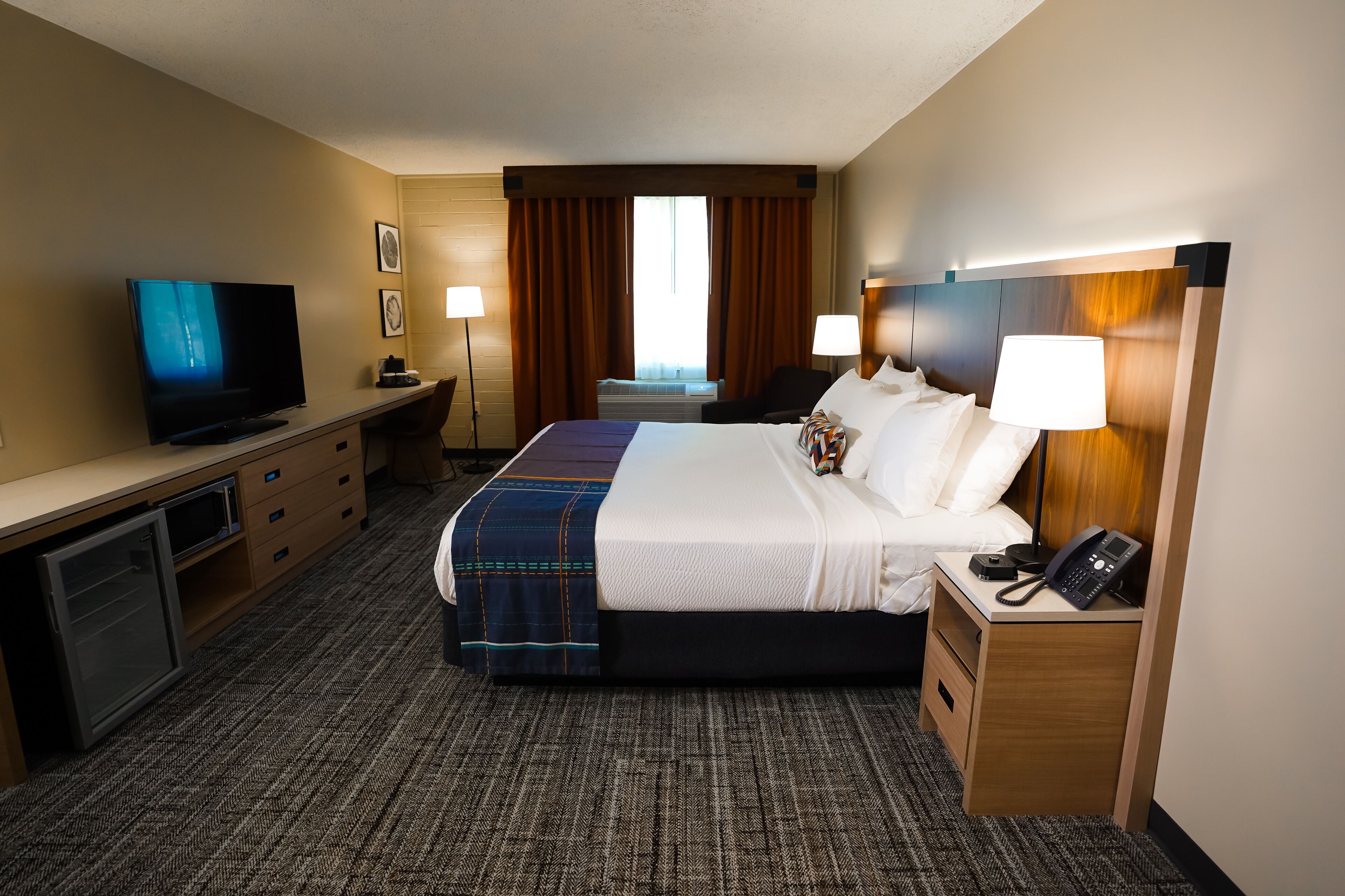 Een hotelkamer met een bed, een bureau met een televisie, een bureaulamp, een nachtkastje met een telefoon en een raam met gordijnen.