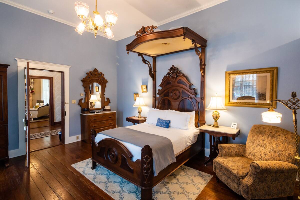 A bedroom with a four-poster bed, a wooden dresser, a chair, and a chandelier.
