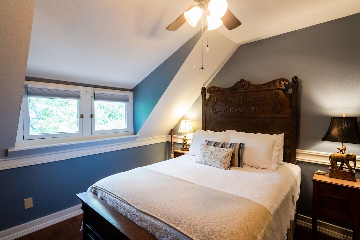 A bedroom with a sloped ceiling, a large bed with a wooden headboard, two bedside tables with lamps, and a window with blinds.