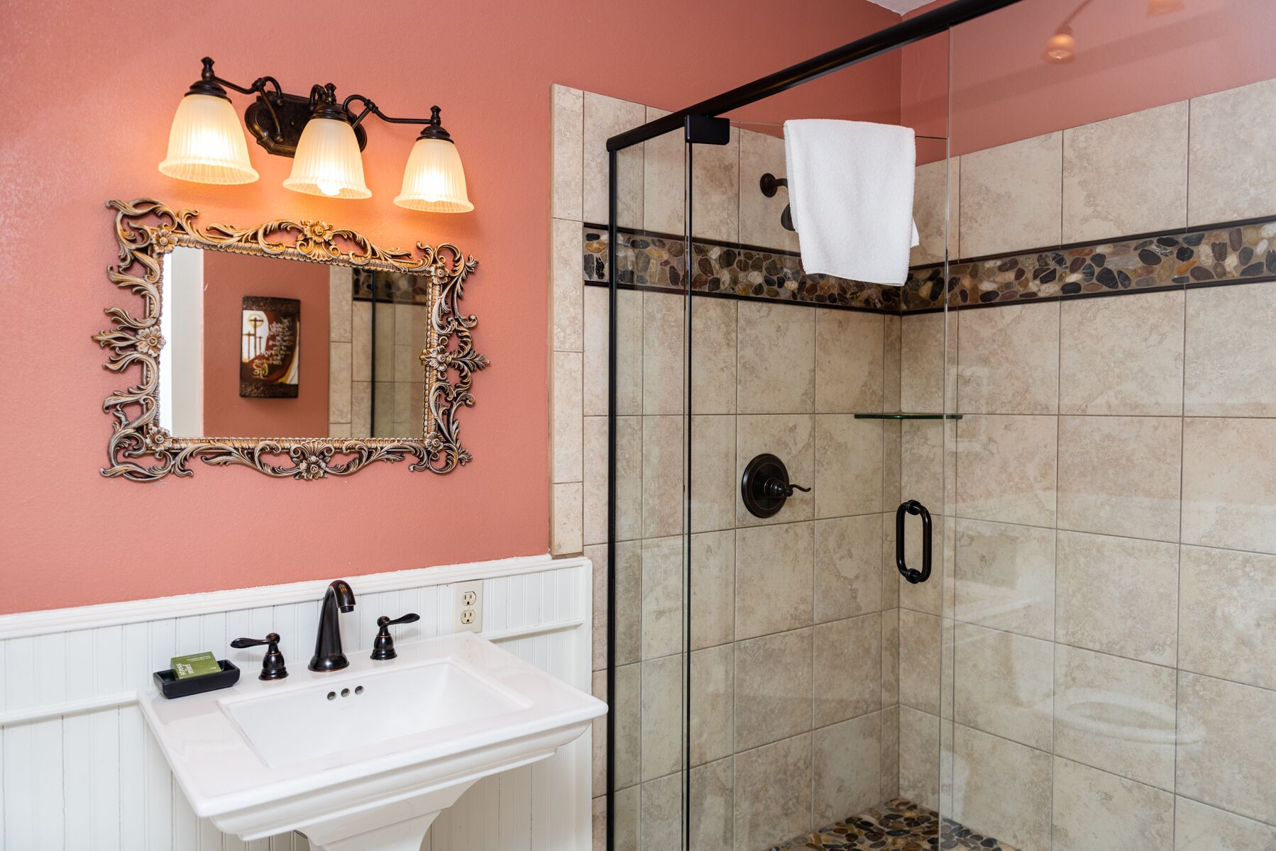 A bathroom with a white sink, ornate mirror, and a shower with a stone tile border.