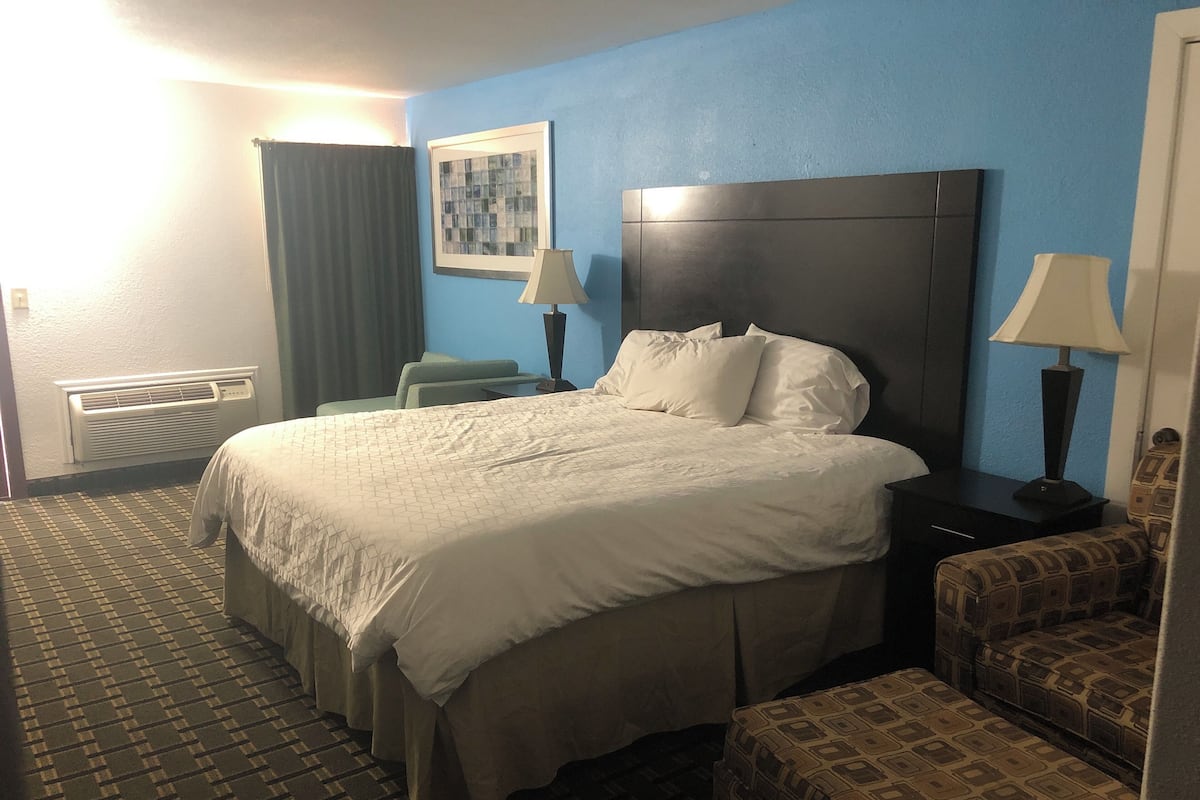 Habitación de hotel con una cama grande, una mesita de noche, una silla, una lámpara, un cuadro en la pared y una ventana con cortinas.