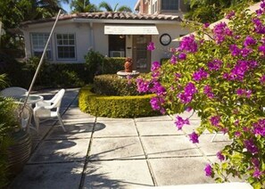 Un patio con una mesa y sillas, un pequeño jardín con una estatua y una casa con techo de tejas.