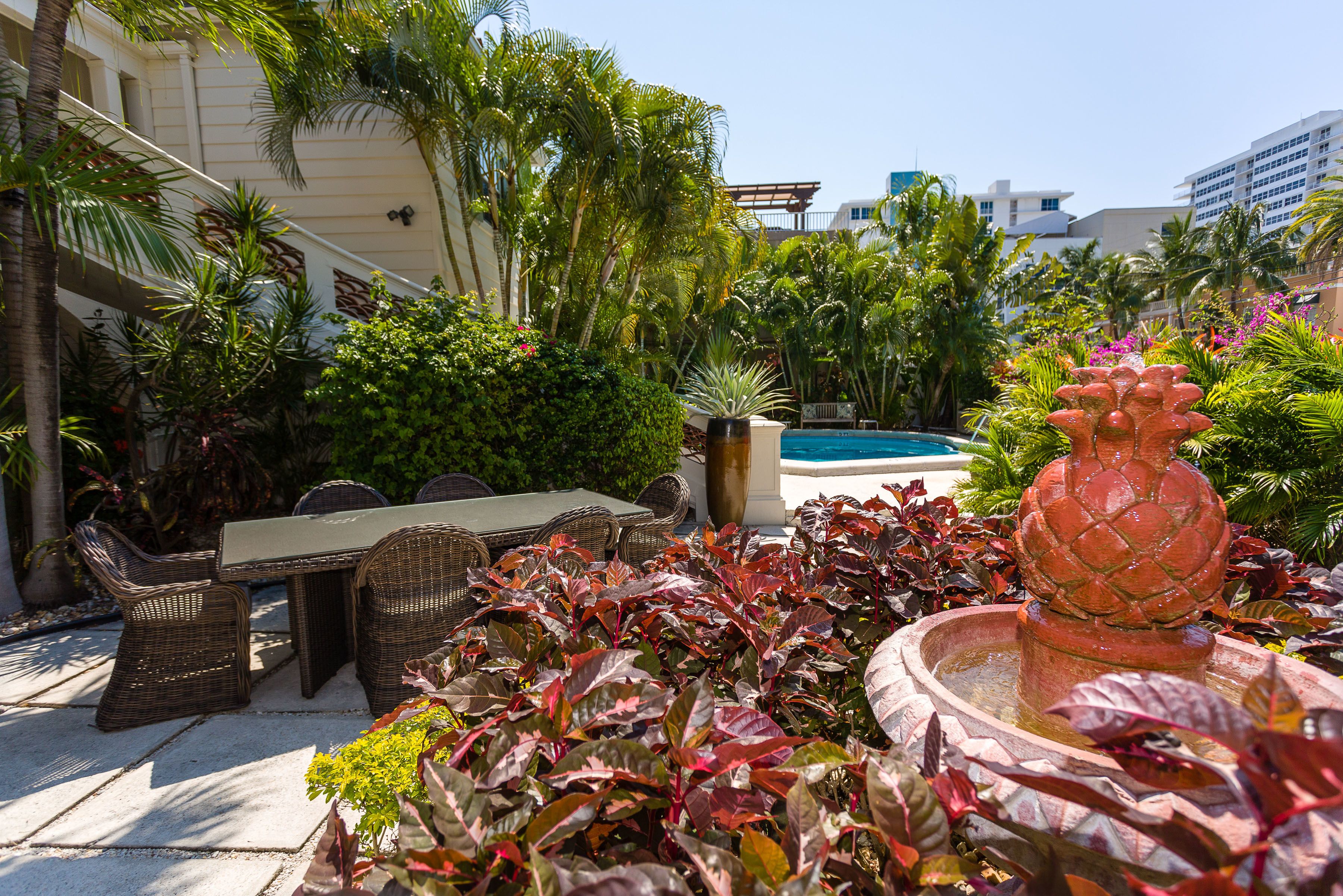 A pool area with a dining table and chairs, surrounded by lush greenery and a decorative fountain.