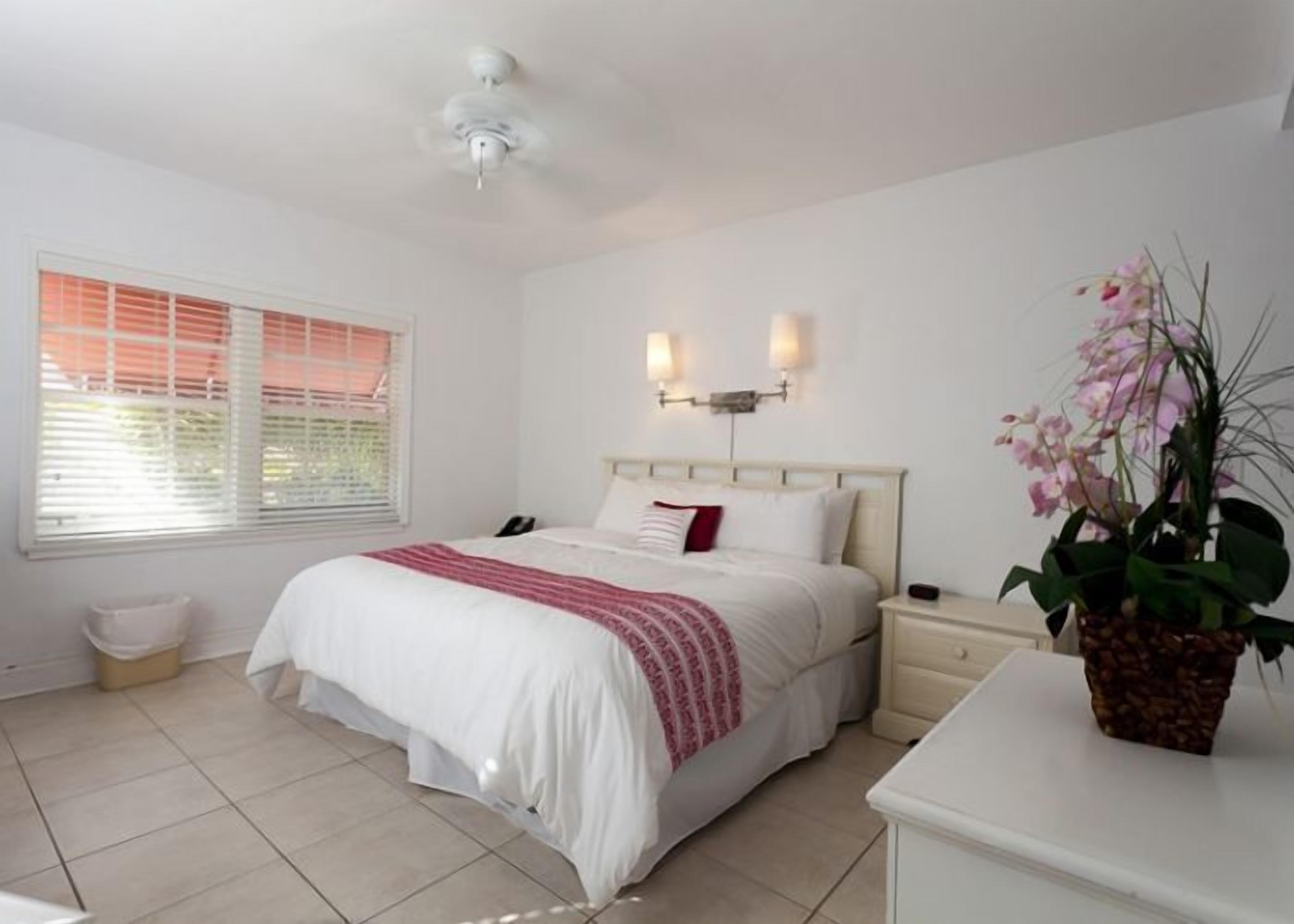 A bedroom with a bed, bedside tables, a ceiling fan, a window with blinds, and a potted plant.