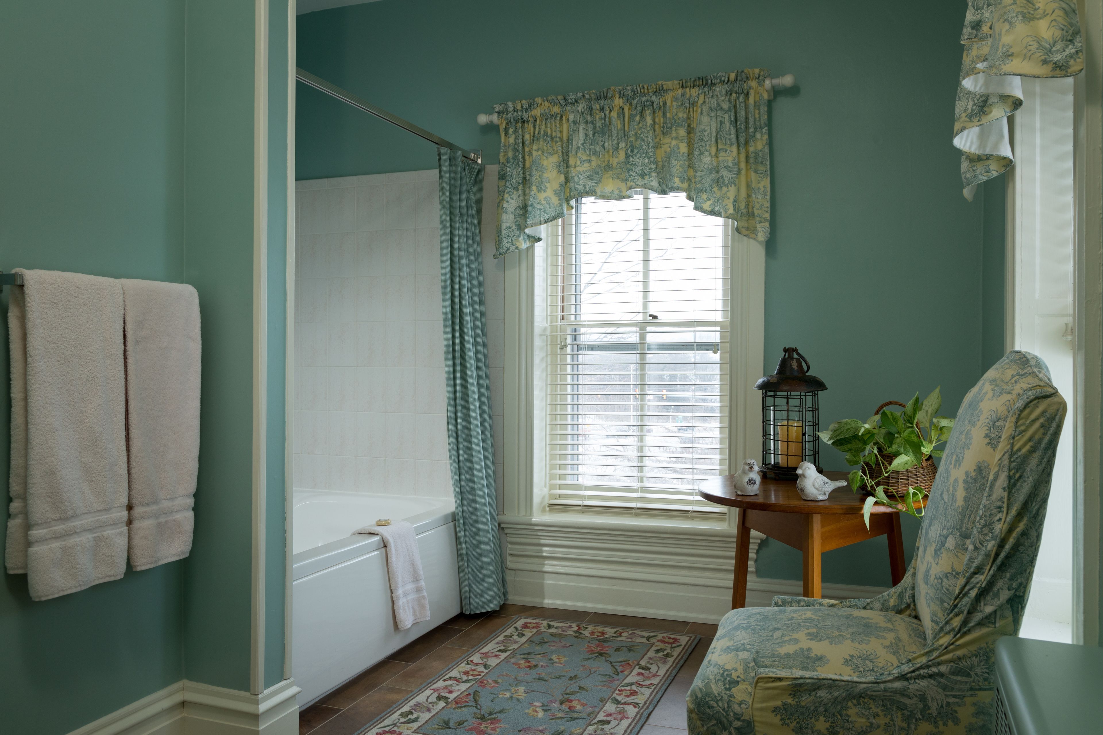Un baño con bañera, una ventana con cortinas, una silla, una mesita con una planta y un perchero.