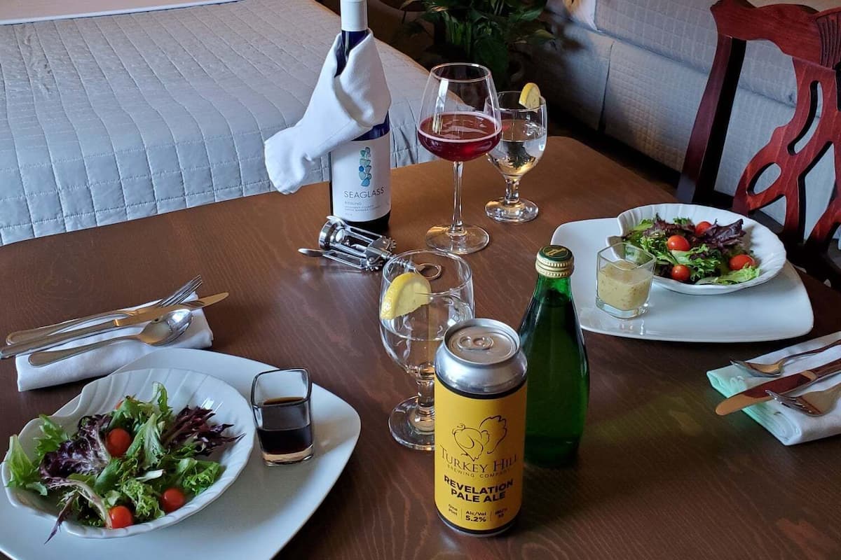 Une chambre d’hôtel avec un lit soigneusement fait, une table à manger dressée avec des assiettes remplies de nourriture, des verres et des bouteilles de boissons.