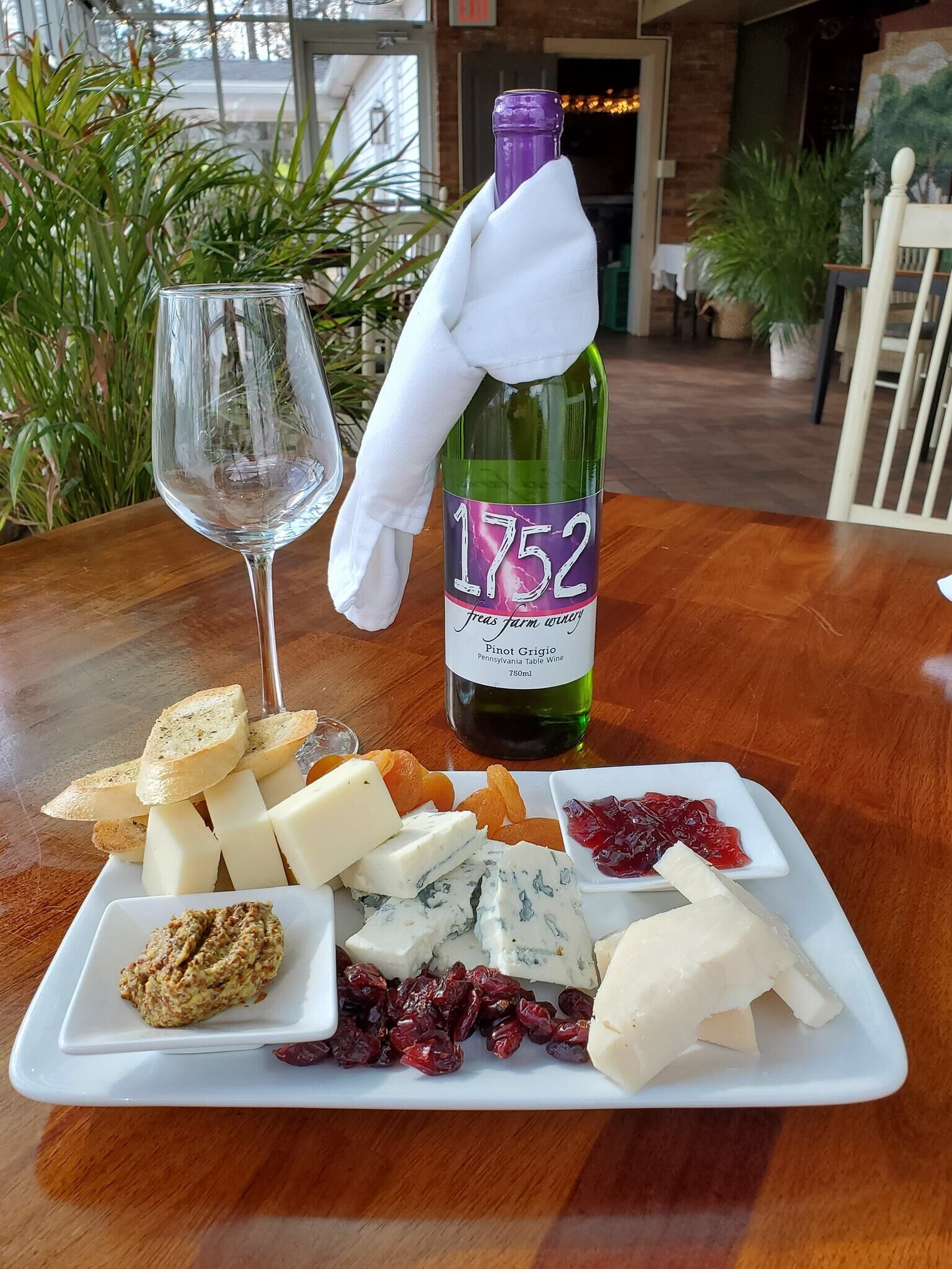 A table with a bottle of wine, a glass, and a cheese board with various cheeses and fruits.