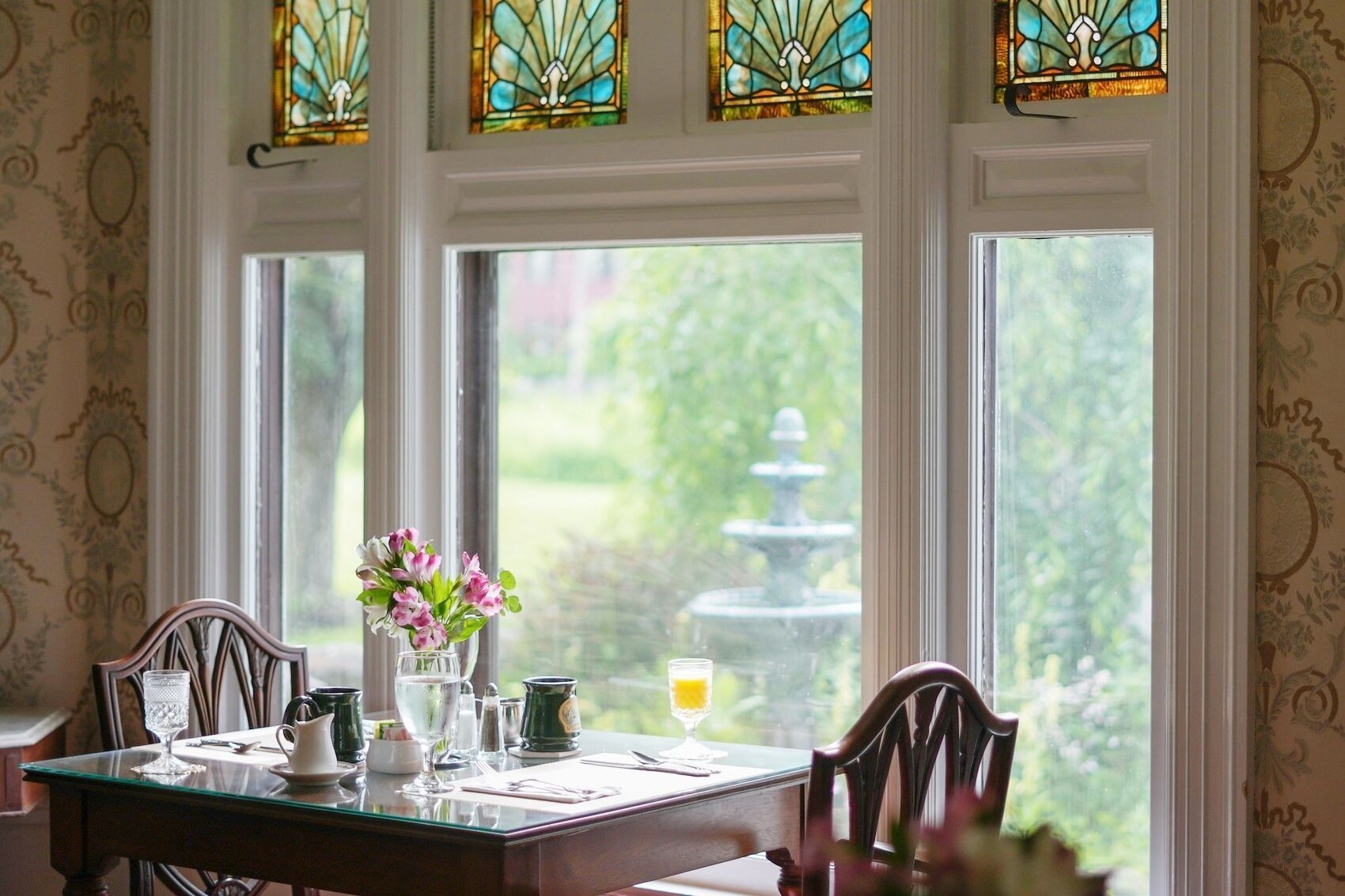 Un comedor con una mesa lista para dos personas, un jarrón de flores y vistas a una fuente a través de amplias ventanas.