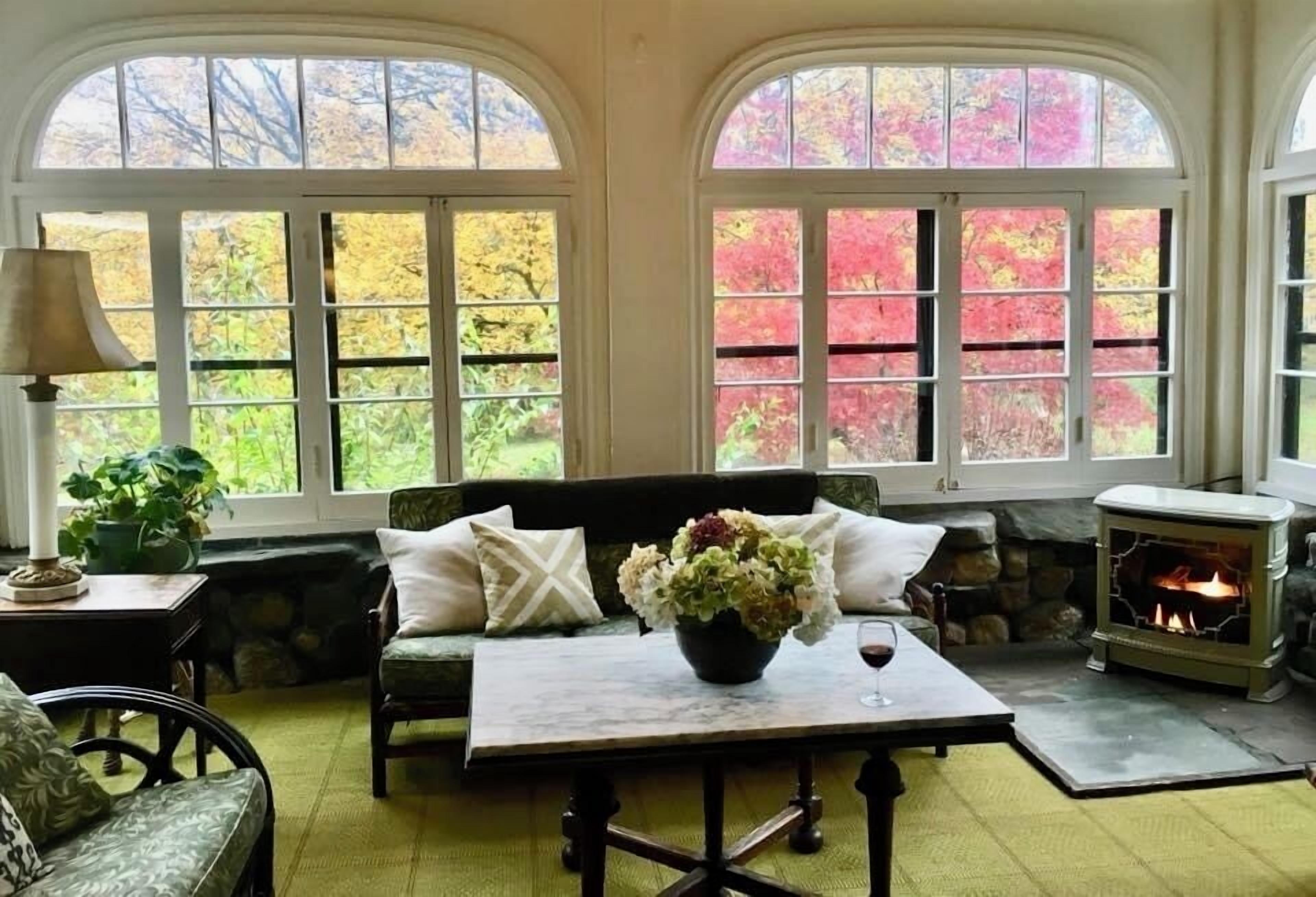 A cozy room with a stone fireplace, a sofa with cushions, a table with a vase and wine glass, and large windows with a view of autumn foliage.