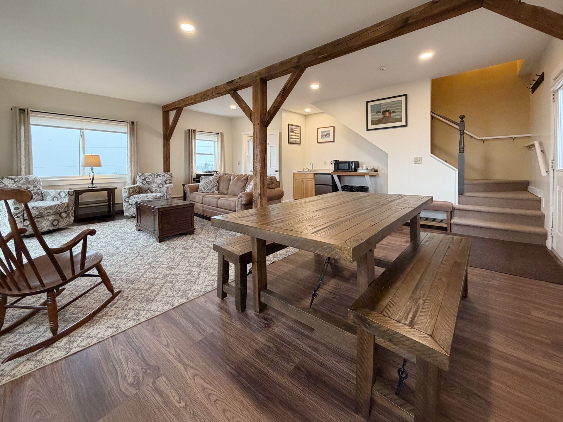 A spacious living room with a wooden dining table, sofas, and a rocking chair. A kitchen is visible in the background.