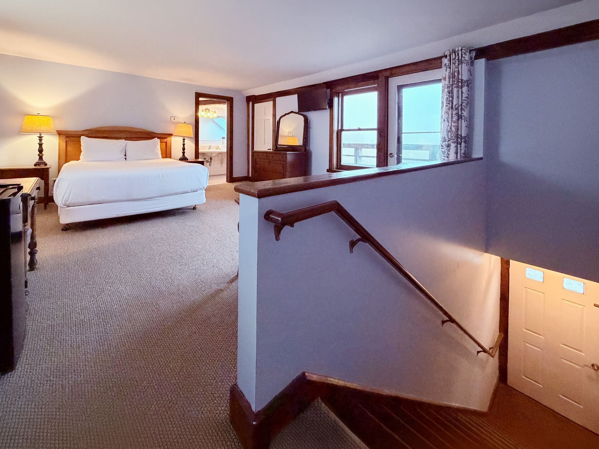 Een hotelkamer met een bed, een bureau, een lamp, een spiegel, een raam met gordijnen en een trap.