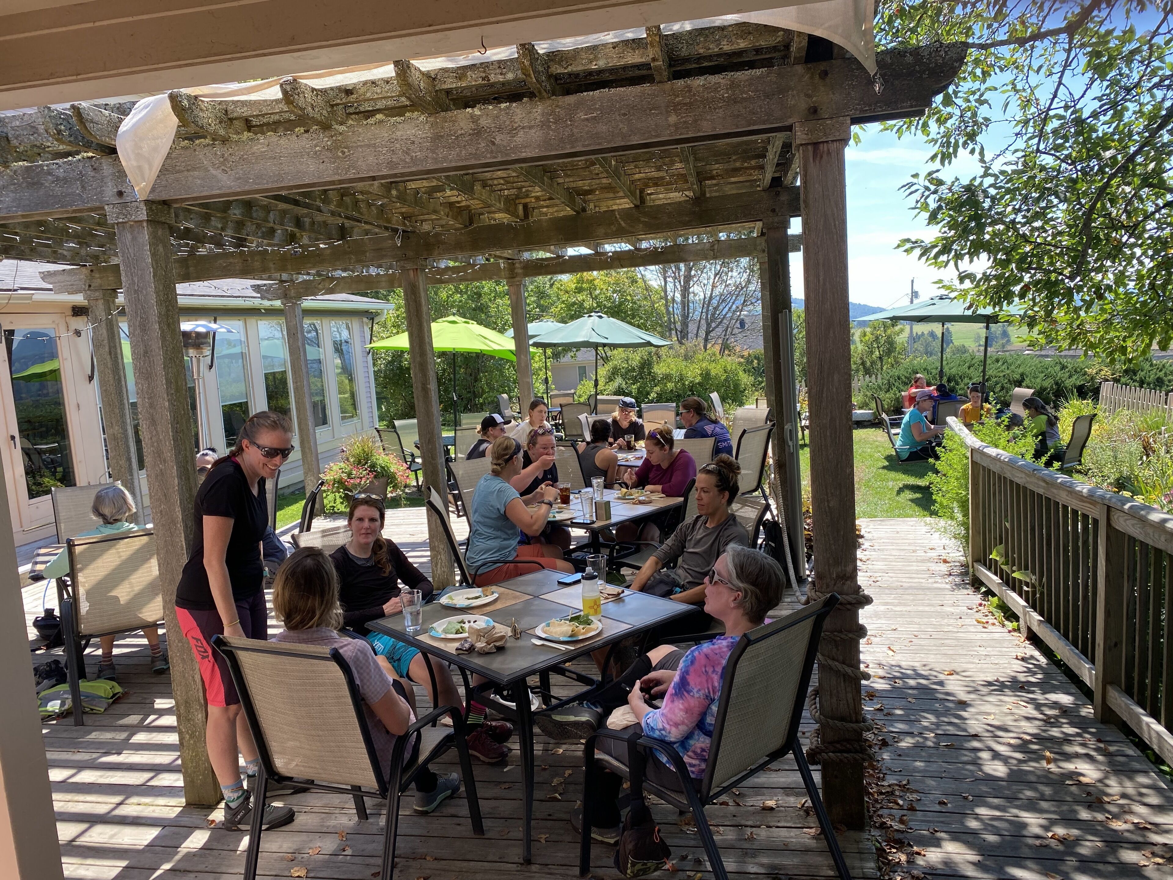 Une terrasse extérieure couverte, dotée de tables et de chaises, et entourée d’une clôture en bois et de verdure.
