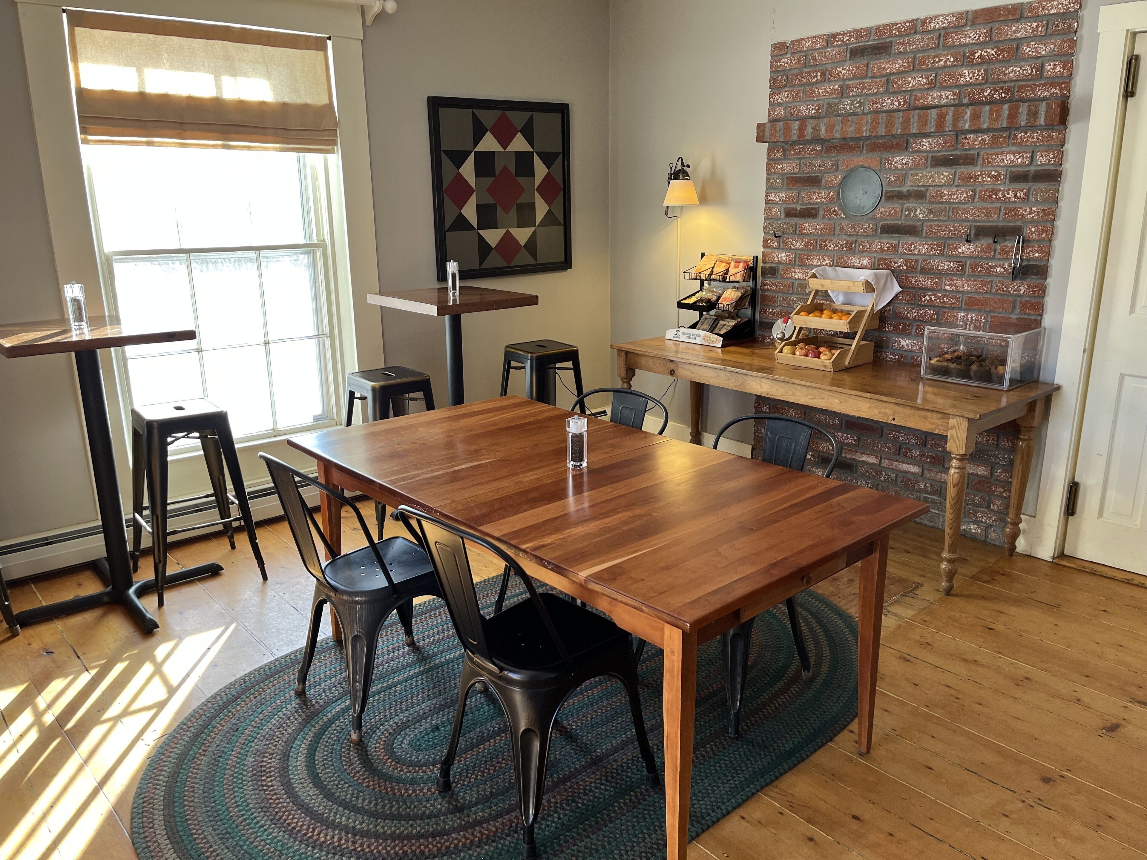 Un coin repas avec une table en bois, des chaises noires, un tapis à motifs, un mur en briques et une fenêtre avec des stores.