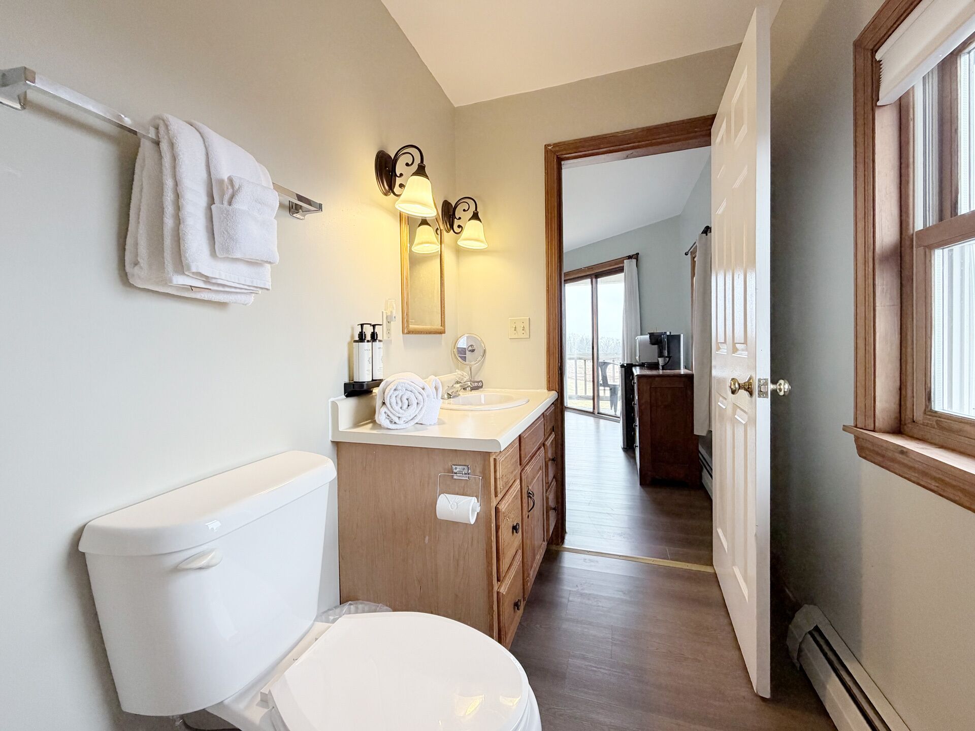 A bathroom with a toilet, sink, and mirror. There is a towel rack with towels, a wooden cabinet, and a door leading to another room.