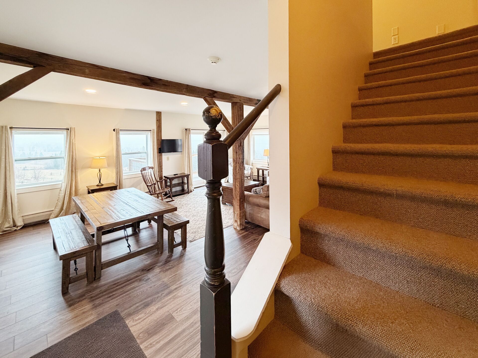 Un escalier avec une rampe en bois et des marches recouvertes de moquette, menant à un espace de vie comprenant une table et des chaises en bois.