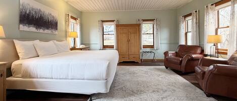 A bedroom with a large bed, two nightstands, a wooden wardrobe, a leather armchair, and a window with curtains.
