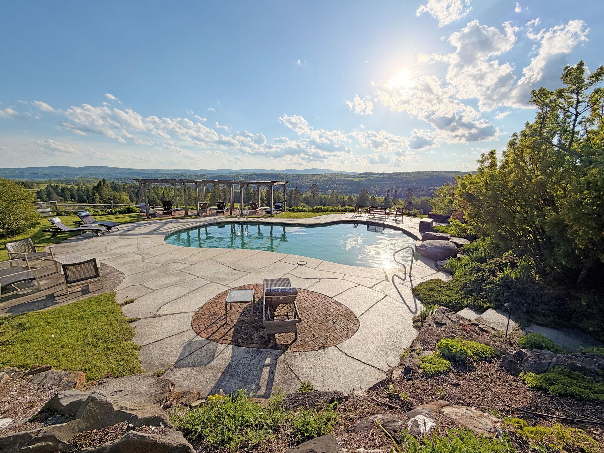 Une piscine extérieure circulaire, dotée de chaises longues et d’une pergola, entourée d’un jardin aménagé et offrant une vue sur des collines lointaines.