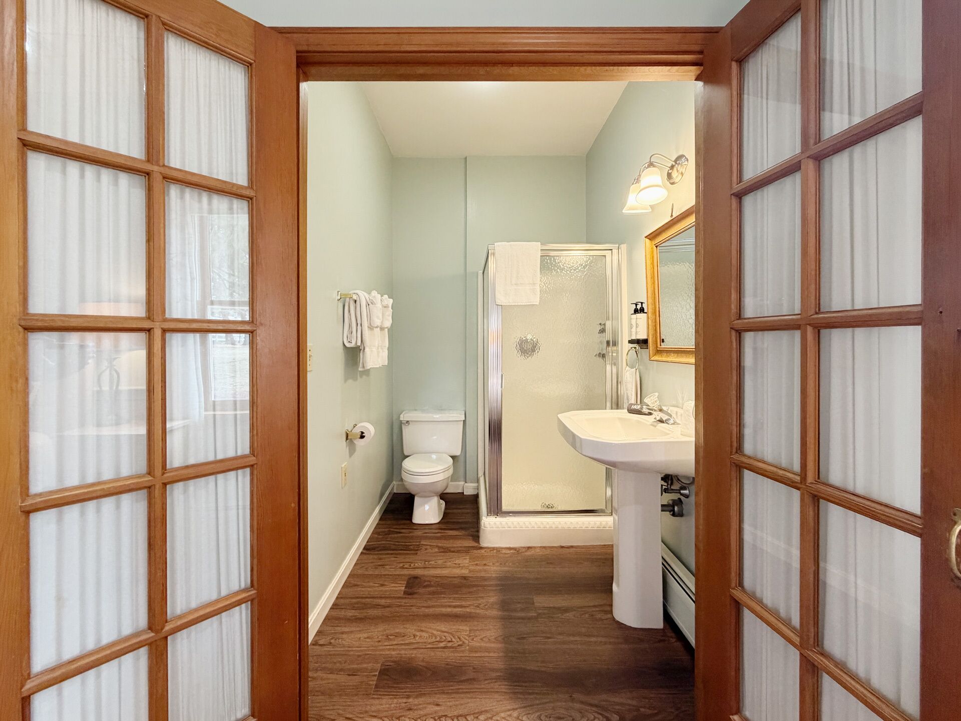 A bathroom with a toilet, shower, and sink, wooden flooring, and frosted glass doors.
