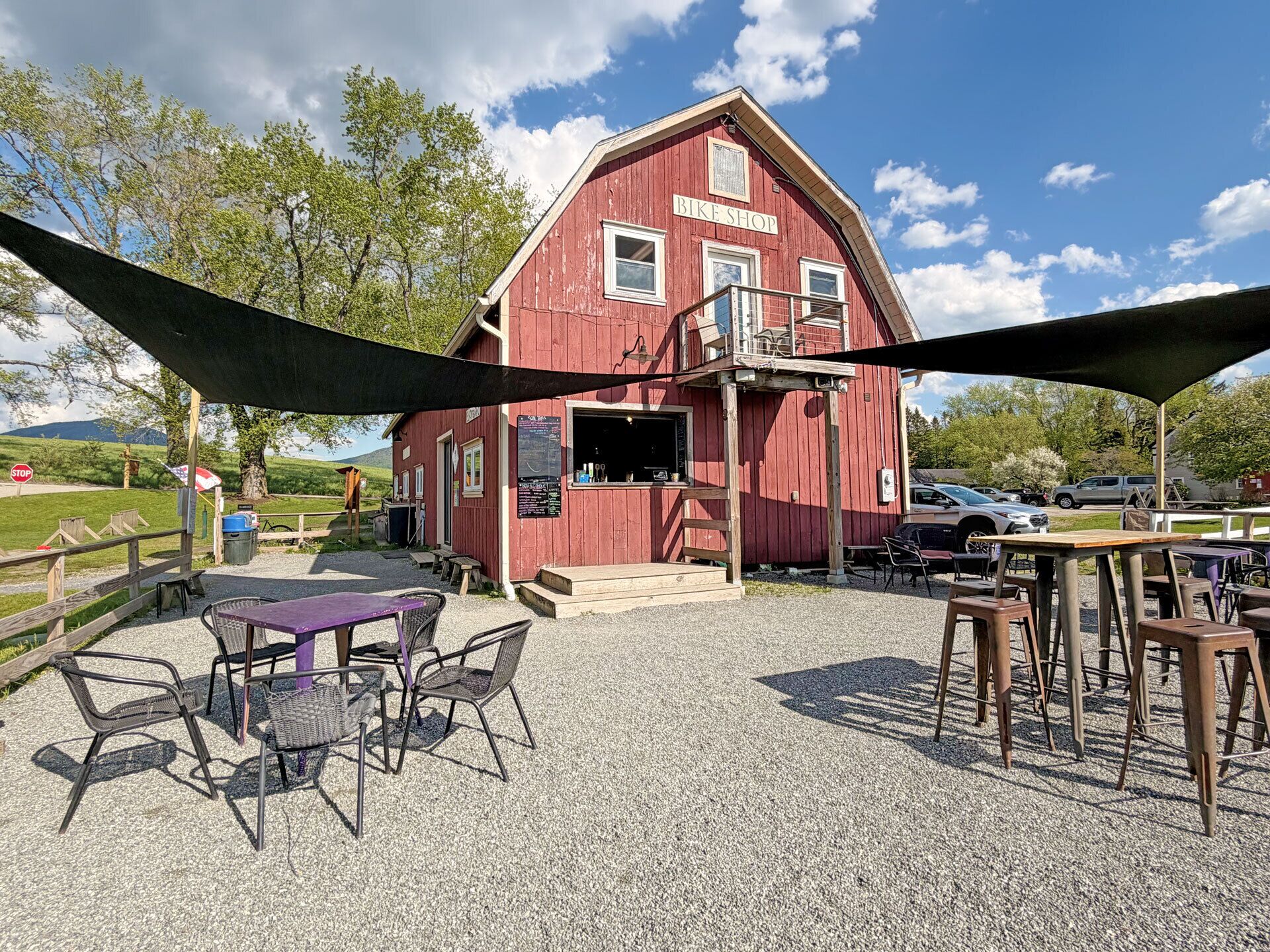 A red building with a sign reading 'BIKE SHOP' and a gravel patio with tables and chairs.