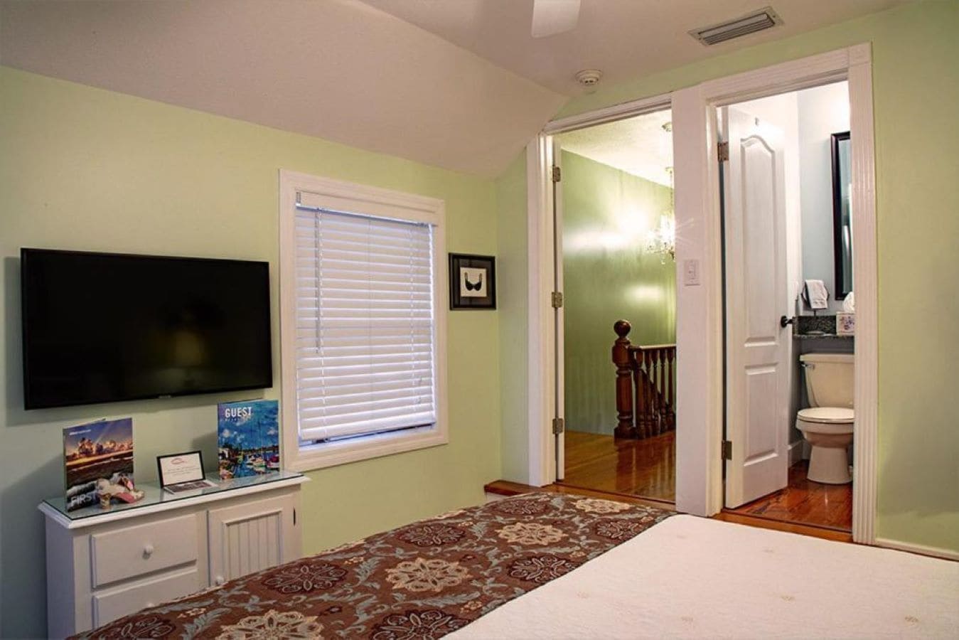 A bedroom with a bed, a television, a window with blinds, a dresser, and a door leading to a bathroom.