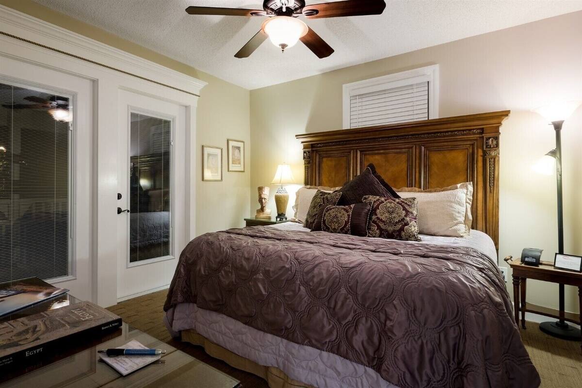 A bedroom with a large bed, a ceiling fan, a nightstand with a lamp, and a door with blinds.