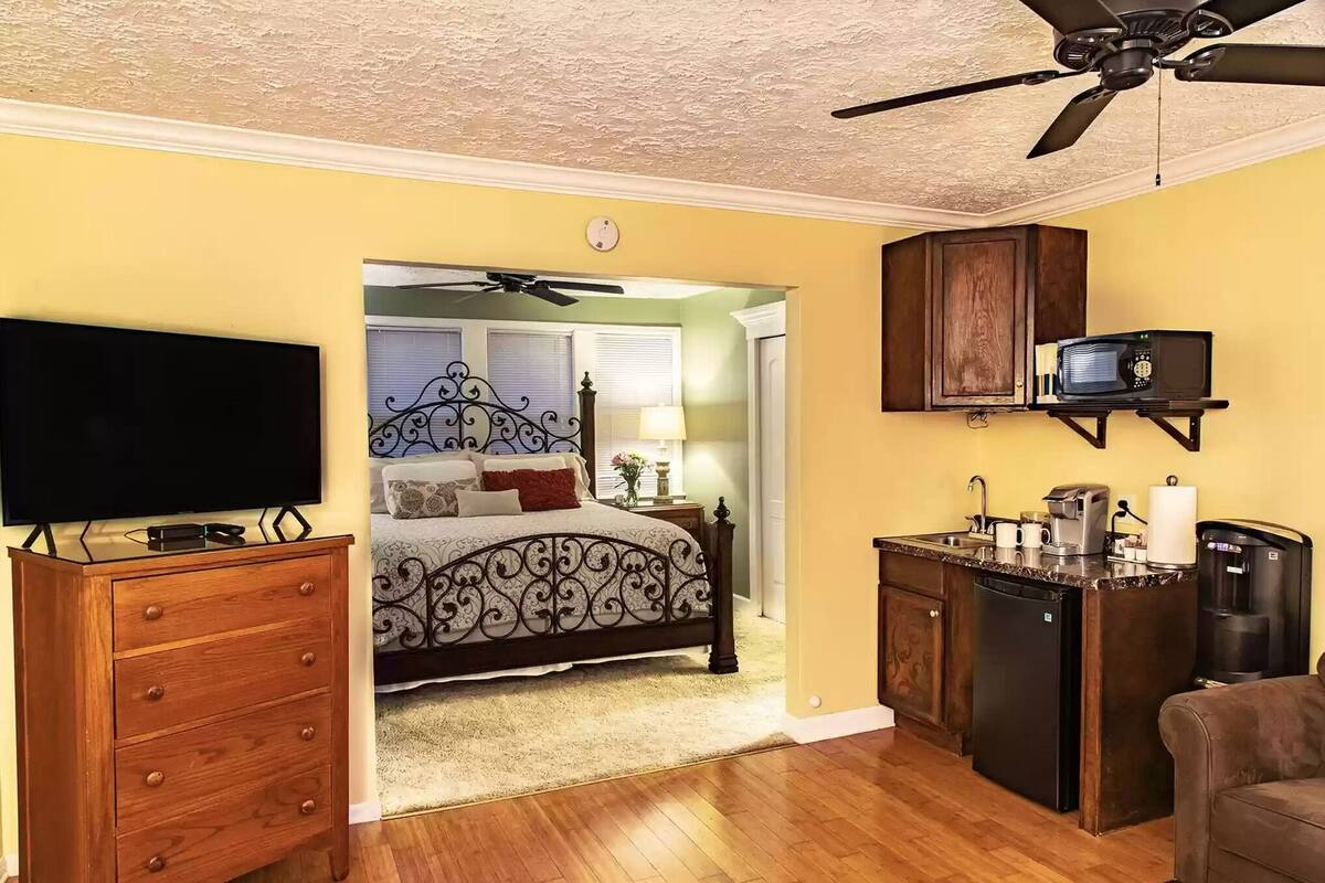 A bedroom with a bed, dresser, TV, ceiling fan, and a kitchenette with a microwave and coffee maker.