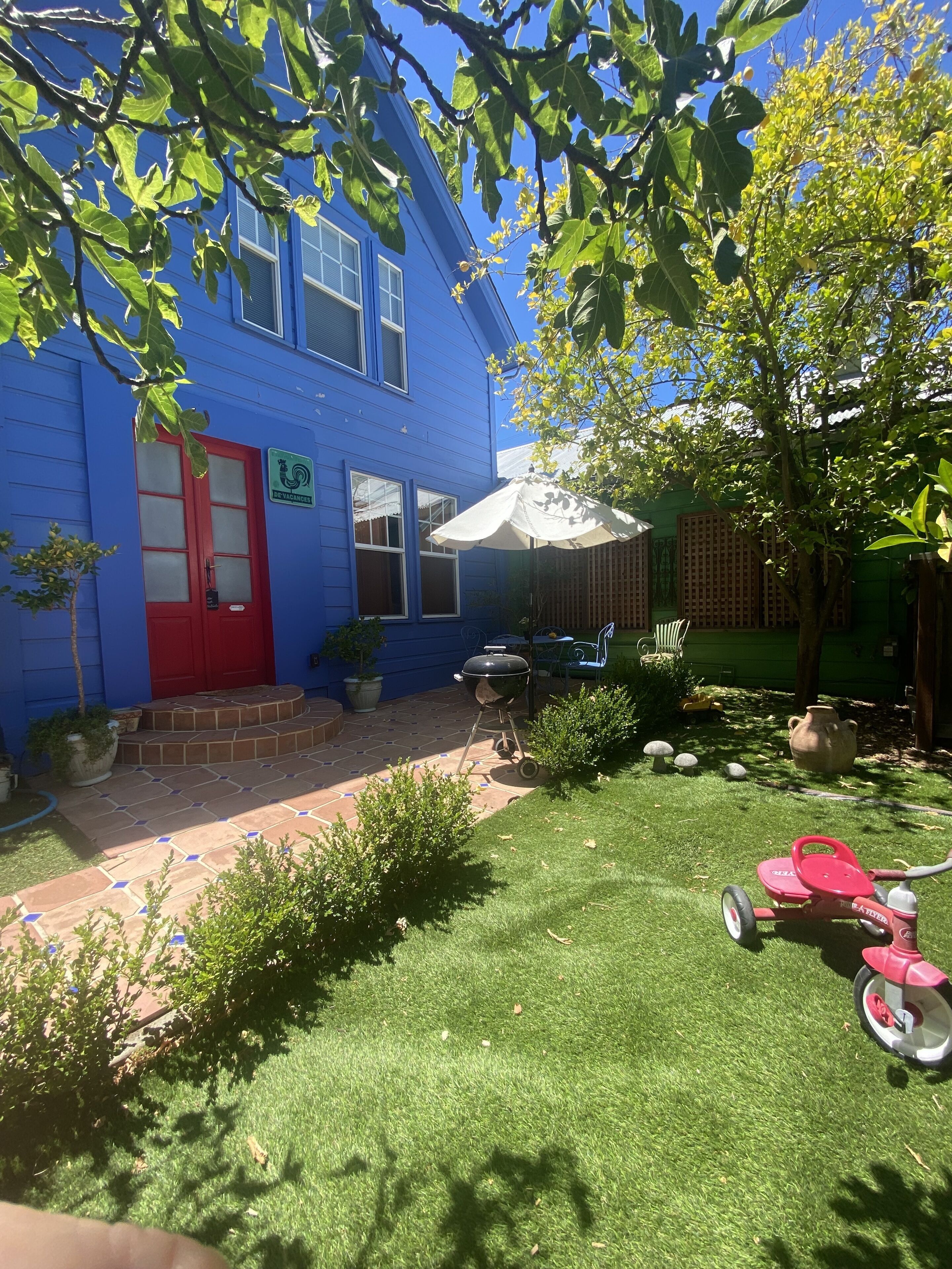 Una casa azul con puerta roja, una parrilla para barbacoa, una triciclo infantil rosa y un patio con plantas en macetas.