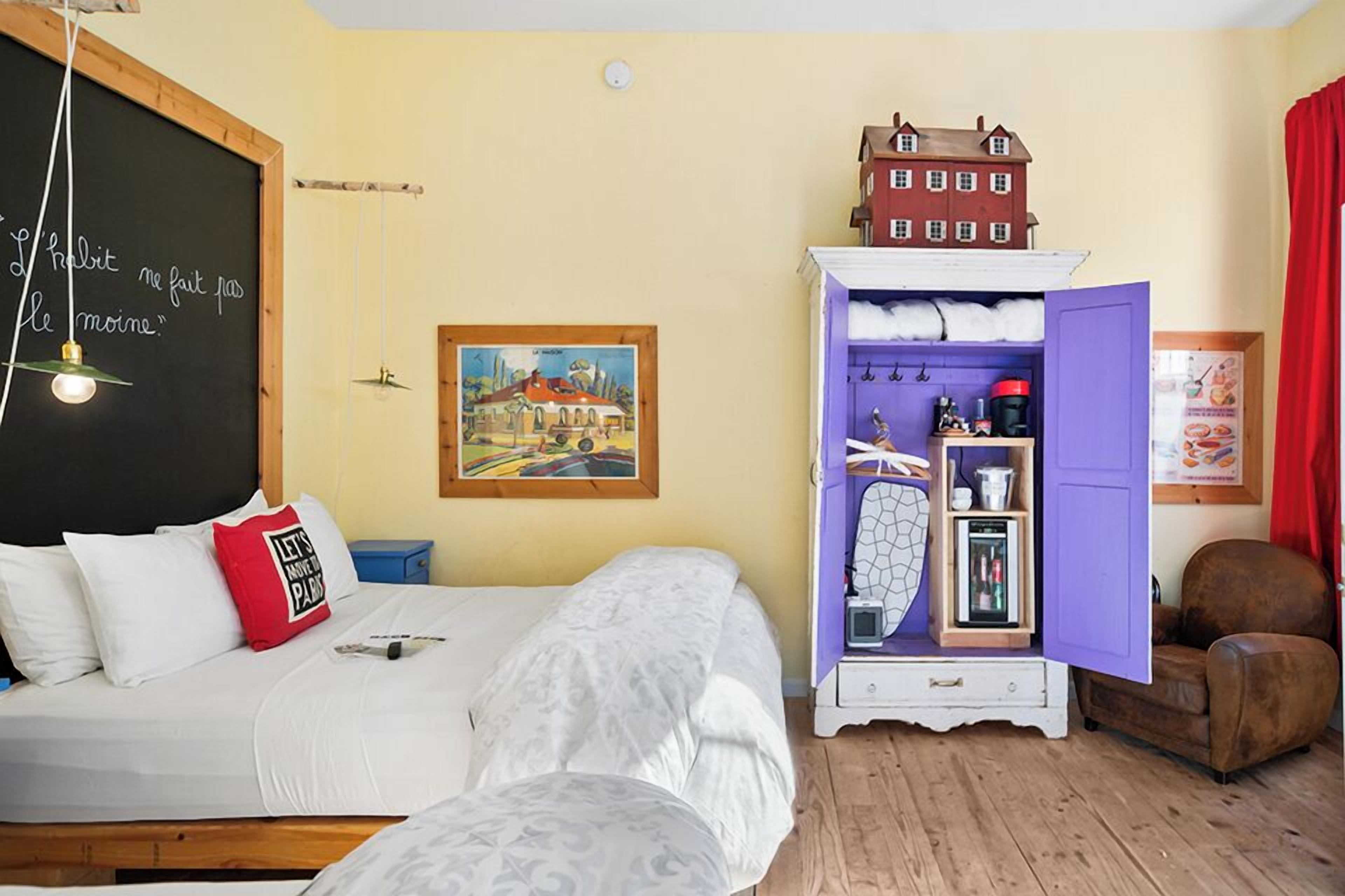 A bedroom with a bed, a dresser with open doors, a small table, and a wall with a chalkboard.