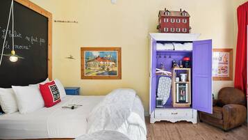 A bedroom with a bed, a dresser with open doors, a small table, and a wall with a chalkboard.