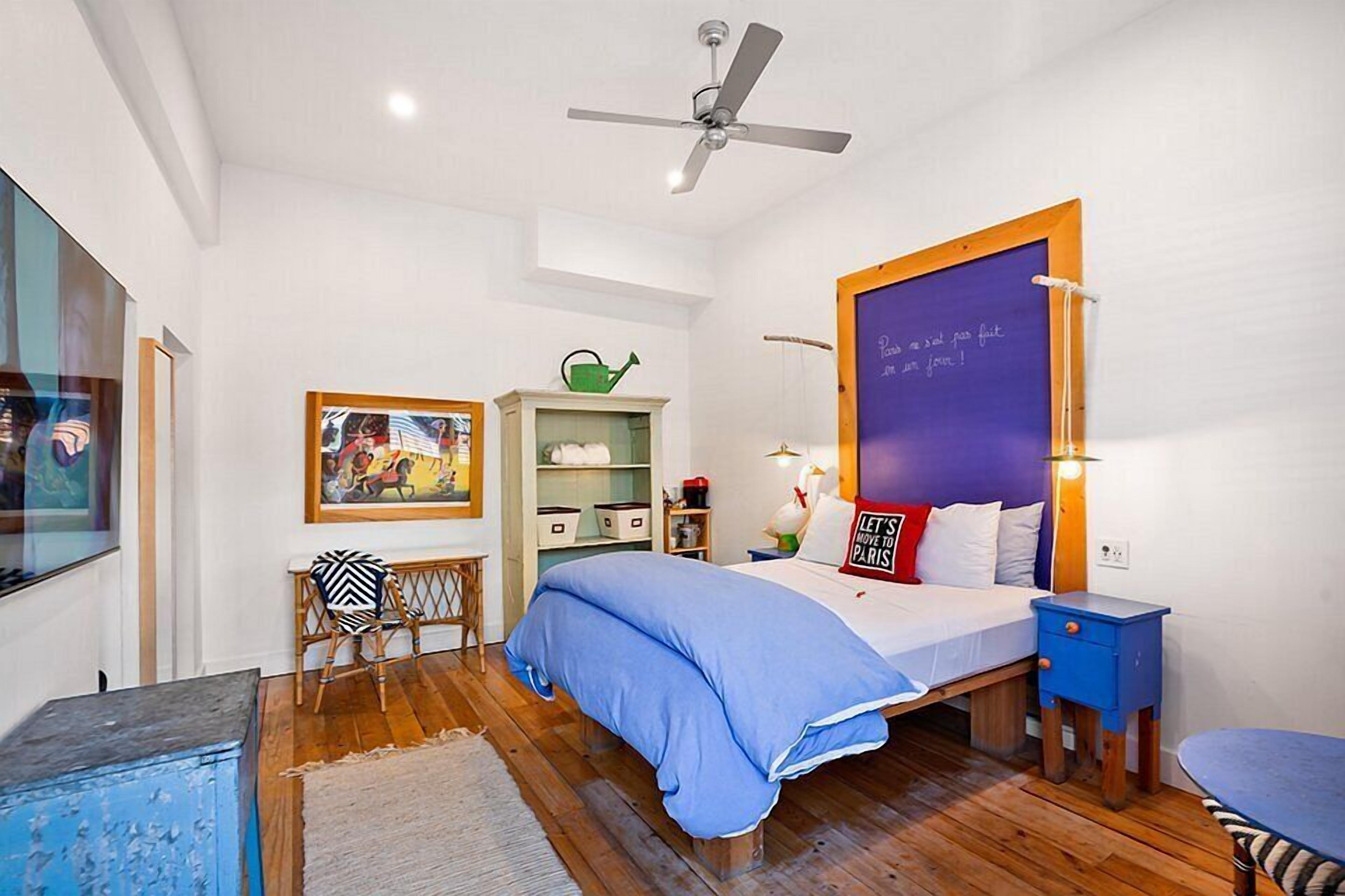 Quarto com cama, ventoinha no teto, penteadeira, cadeira, mesa e parede com quadro negro.