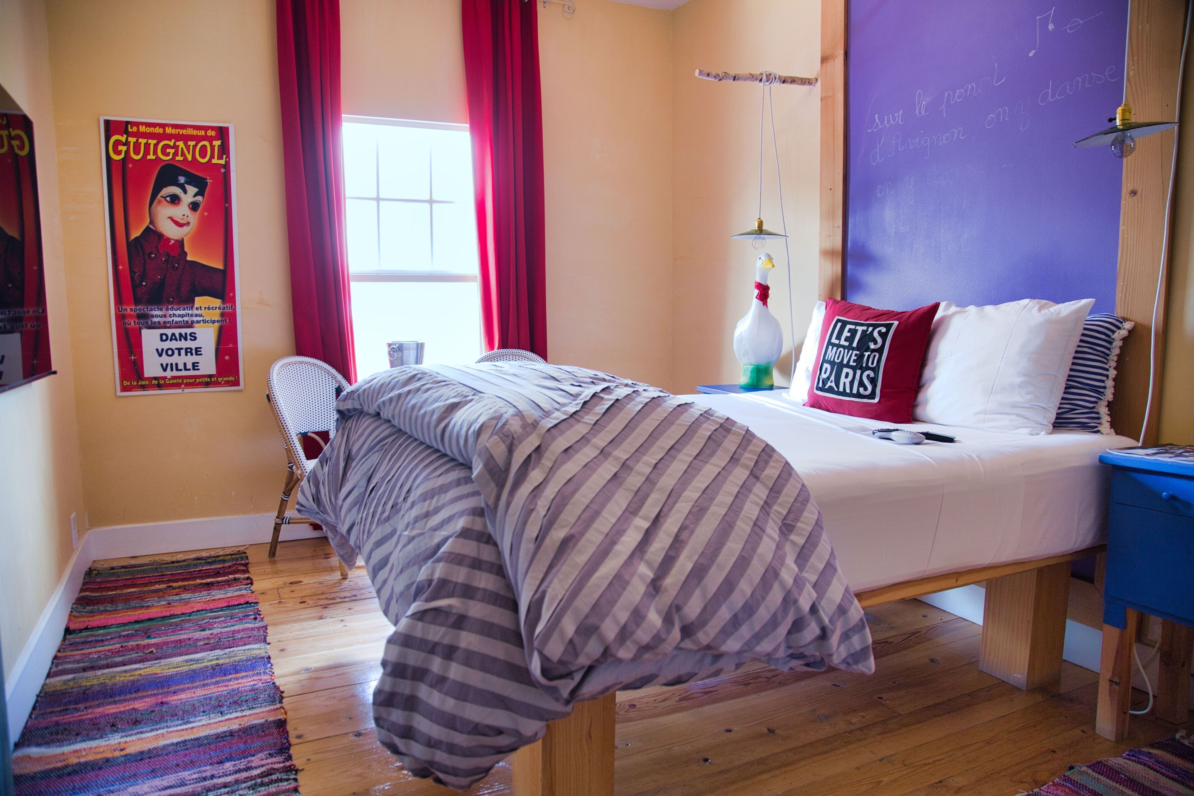 Un dormitorio con cama, una silla, un ventanal con cortinas rojas, un cartel en la pared y una mesita de noche azul.