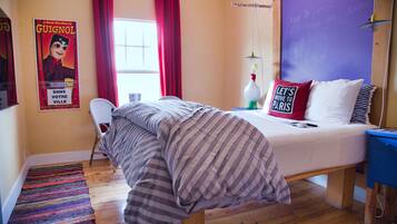 Un dormitorio con cama, una silla, un ventanal con cortinas rojas, un cartel en la pared y una mesita de noche azul.