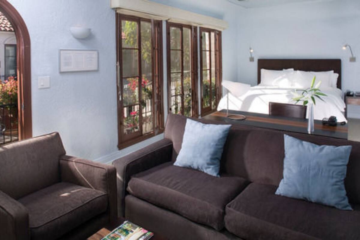 A room with a brown sofa, a wooden side table, a bed with white linens, and a window with a view of greenery.