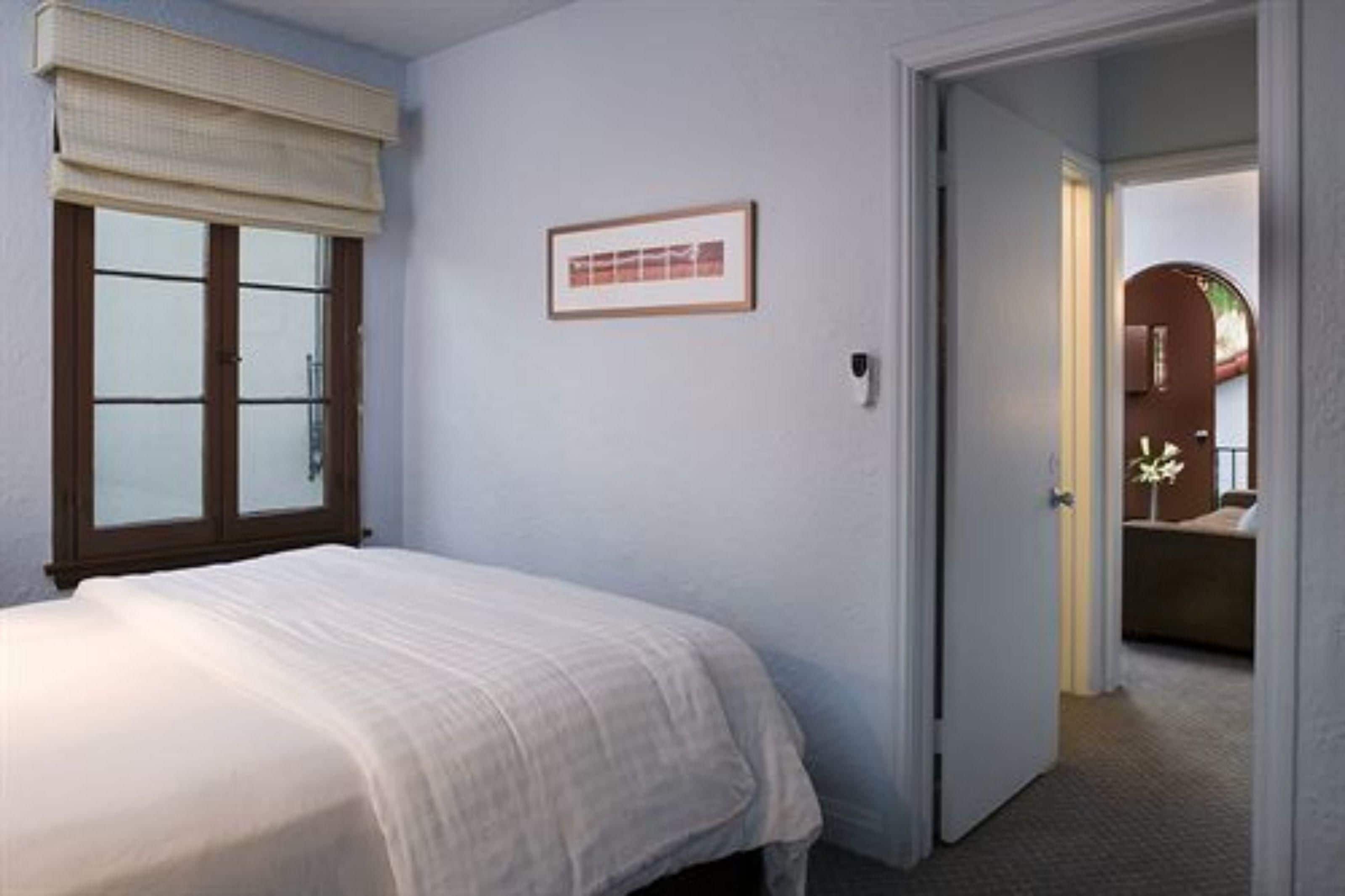 Un dormitorio con cama, ventana con persianas, un cuadro en la pared y una puerta que da a otra habitación.