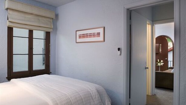 Une chambre à coucher avec un lit, une fenêtre avec des stores, un tableau encadré au mur et une porte donnant sur une autre pièce.