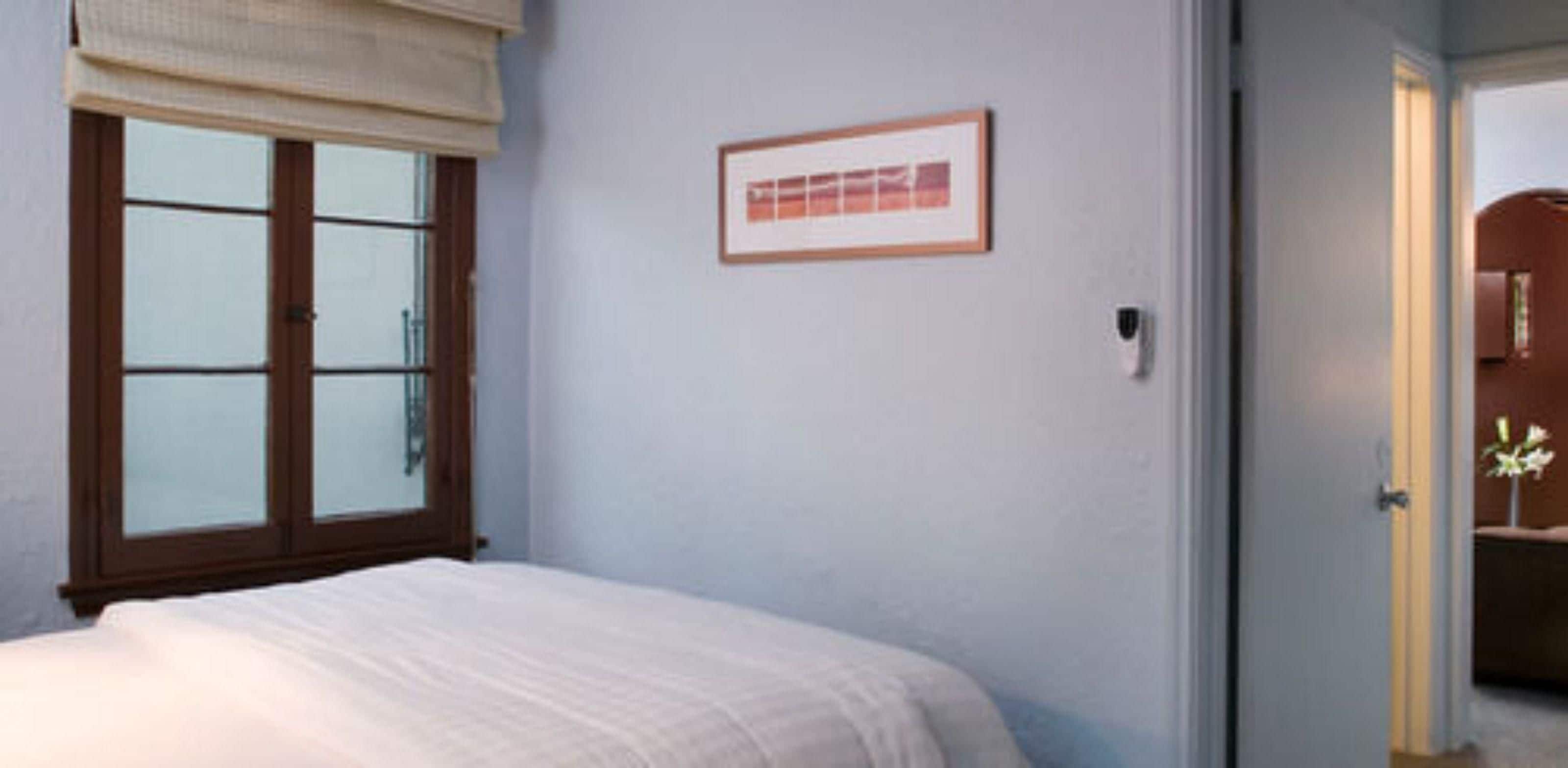 A bedroom with a bed, a window with blinds, a framed picture on the wall, and a door leading to another room.