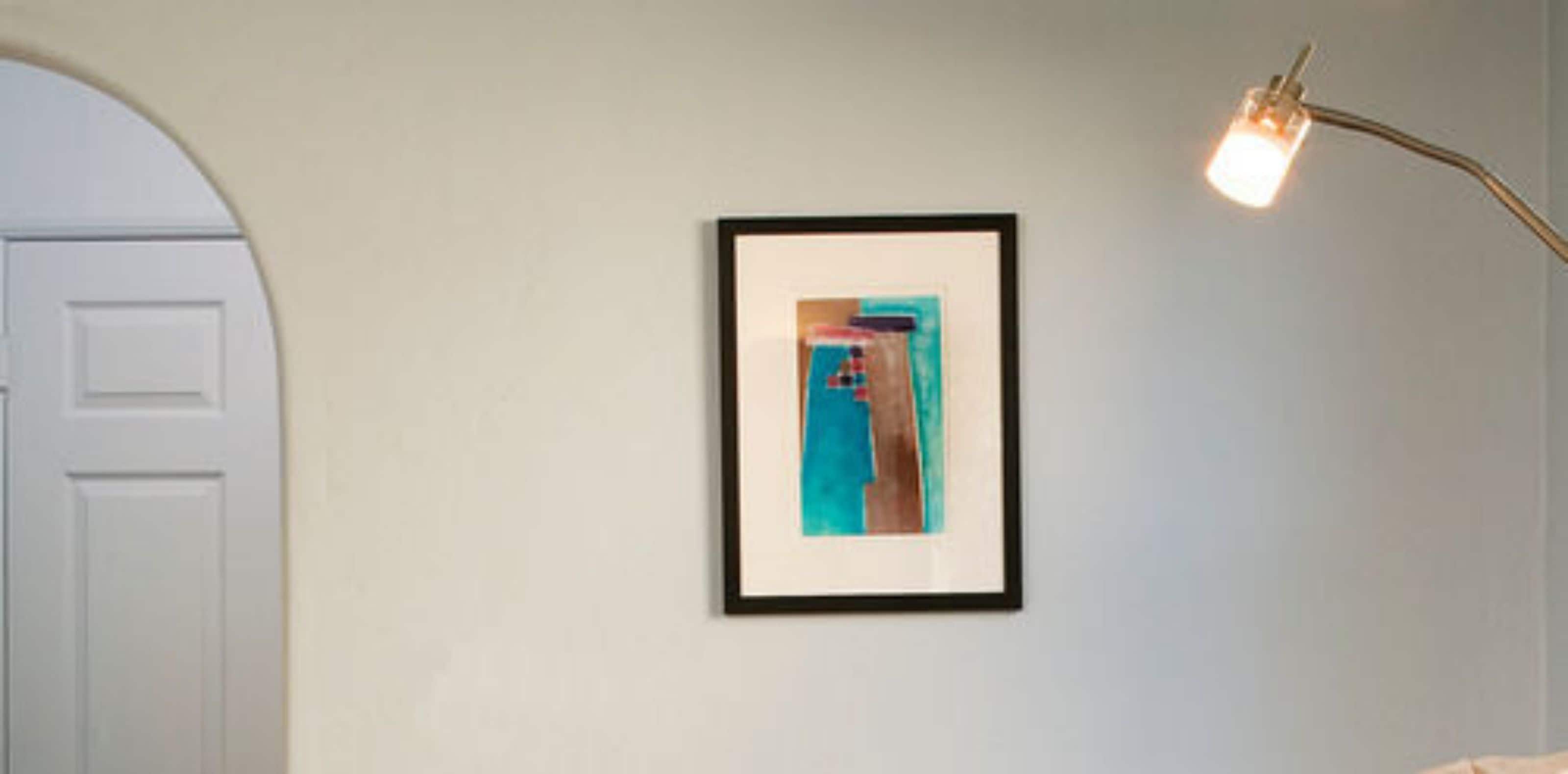 A framed abstract painting on a plain wall, a white door, and a floor lamp with a light bulb.